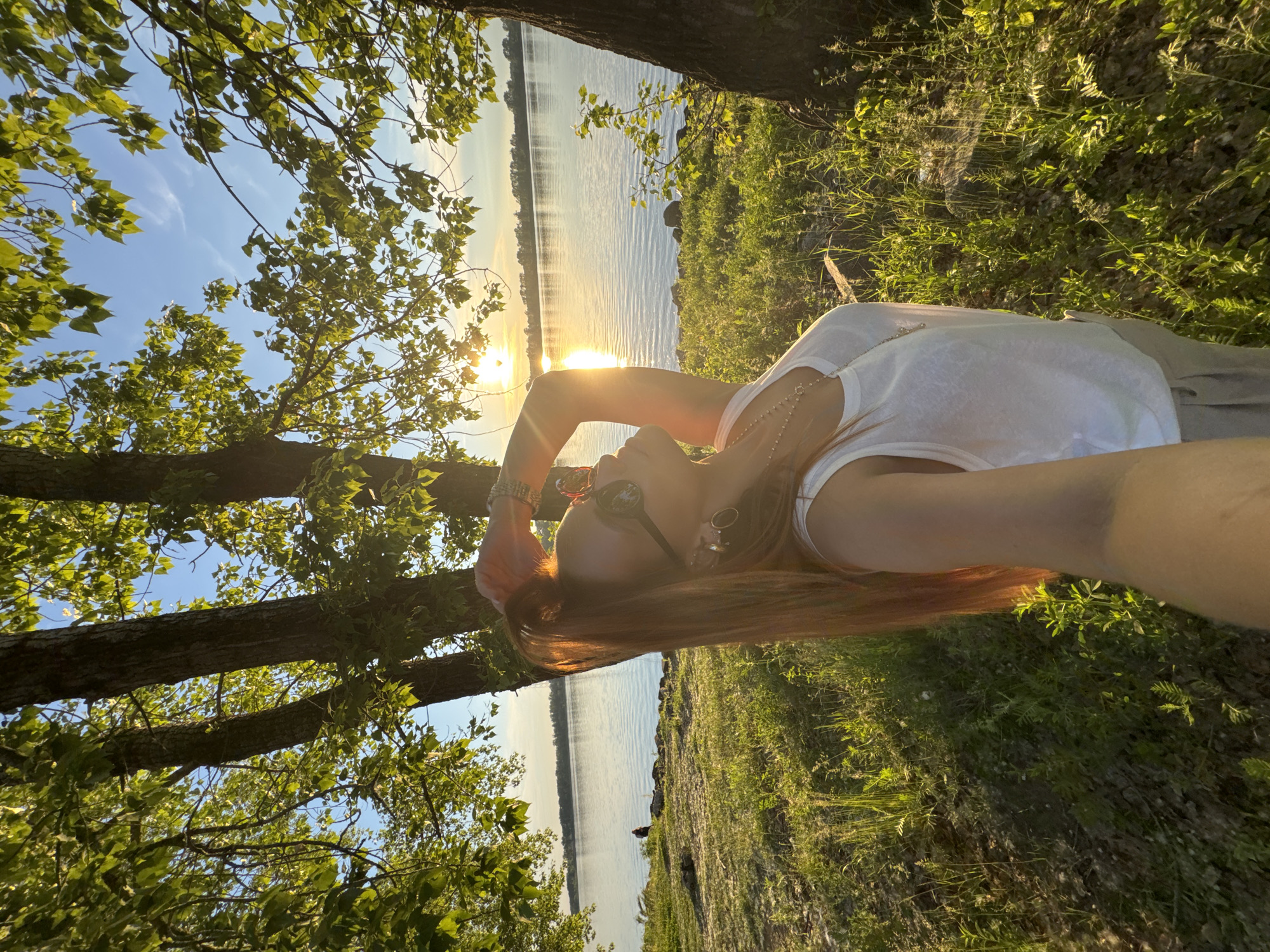 An overhead shot captures a woman with long, auburn hair standing by a river at sunset. She is wearing a white tank top, light-colored pants, and sunglasses, with her right arm raised to her head. The sun is setting over the water, casting a golden glow on the scene and creating a lens flare. Trees with green leaves frame the top of the image. The riverbank is covered in grass and small stones. In the distance, a single person can be seen walking near the water's edge. The location is the Kama River embankment in Chaikovsky, Russia.