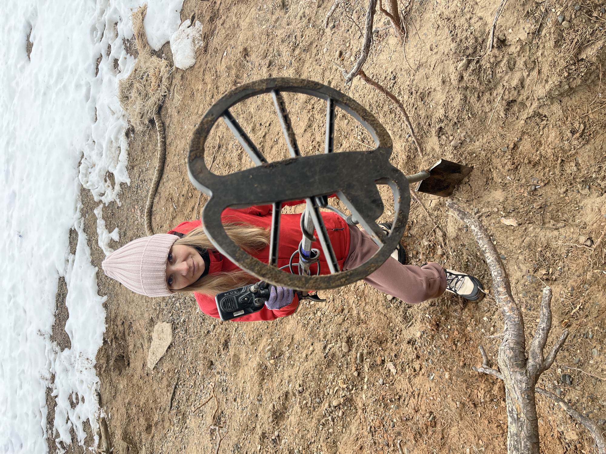 A young woman in a pink beanie, red jacket, and mauve pants holds a metal detector in a dirt and snow environment. She is smiling at the camera. To her right is a small shovel. The surrounding ground is covered in dirt, gravel, and tree branches. A patch of snow sits adjacent to the dirt. The location is possibly Chaikovsky, Russia.