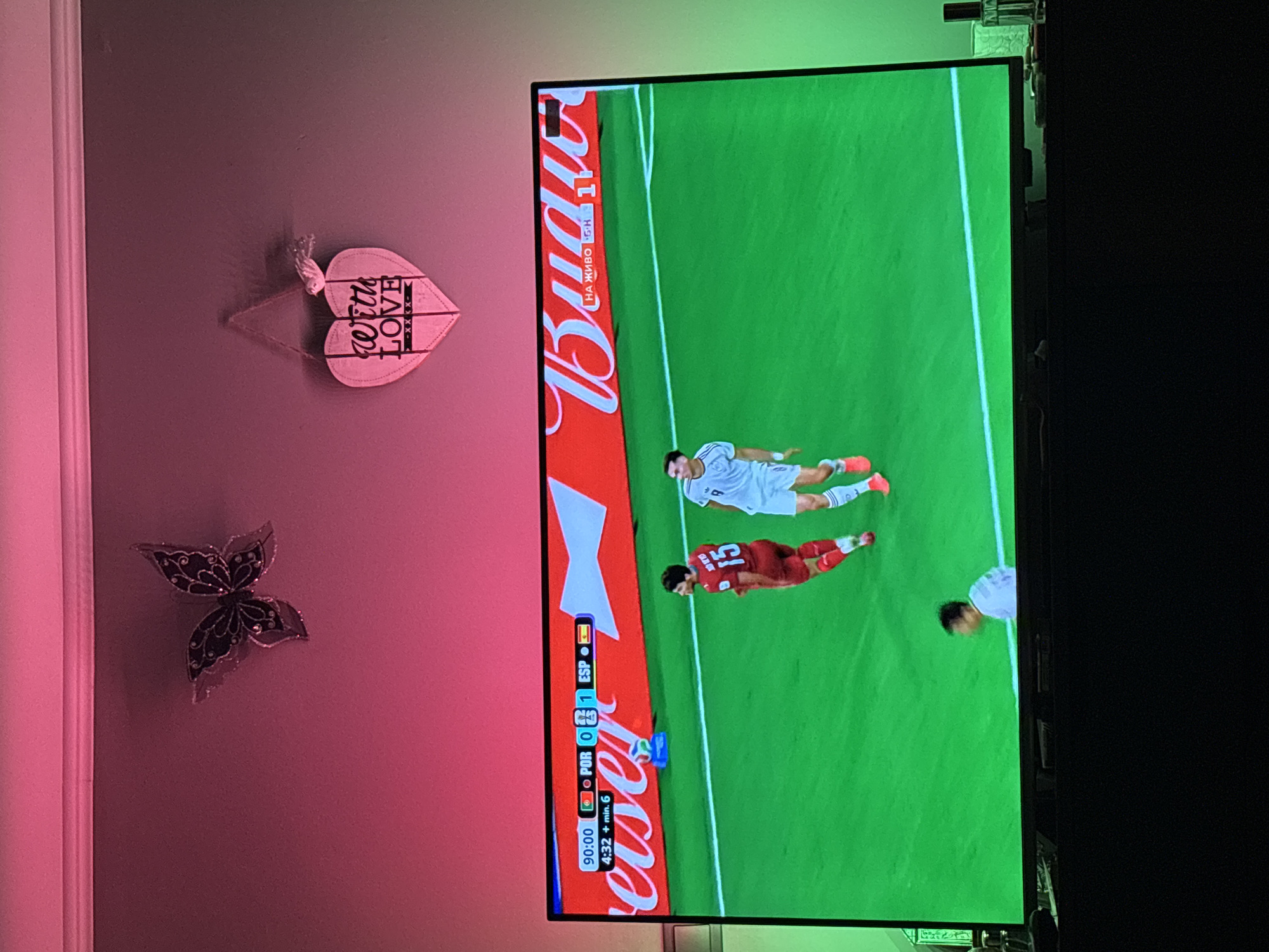 The image depicts a television screen displaying a soccer match. The score shows Portugal with 0 goals and Spain with 1 goal at 90:00, with an additional 4:32 minutes of stoppage time. Two players are visible on the green field. One player is wearing a red jersey with the number 15. The other player is wearing a white jersey. A large red advertisement with the word "Budweiser" is displayed across the top of the screen, with Russian text that translates to "LIVE BROADCAST". Above the television, on a pink wall, are two decorative items: a black butterfly and a white heart-shaped plaque that reads "with LOVE". The overall scene suggests a living room environment where someone is watching a sporting event. No specific location in Marten, Bulgaria, can be identified from this image.