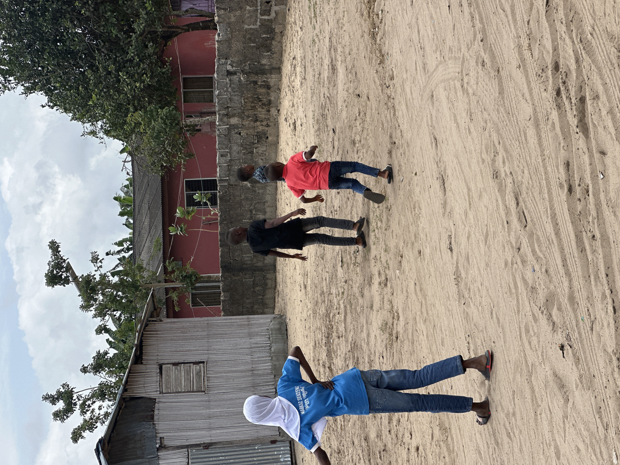 In Badagry, Nigeria, on a sandy lot, four young boys are present. The boy on the left wears a blue shirt with "MARKAZ SEKEENI HAPPY" text, a white hijab, and blue jeans. He faces away from the camera with his hands on his hips. To his right, a boy in a black t-shirt and grey pants walks away from the camera. Another boy in a red t-shirt and blue jeans runs with his arms outstretched, looking to the right. Behind him, another boy in a blue patterned shirt and grey pants walks away. In the background, a weathered, light-colored building with a corrugated metal roof and wooden shutters stands to the left. To the right of that, a pink building with a dark roof and windows is visible, bordered by a low stone wall. Trees with green foliage are scattered throughout the scene. The ground is covered in sand with visible tire tracks and scattered debris.