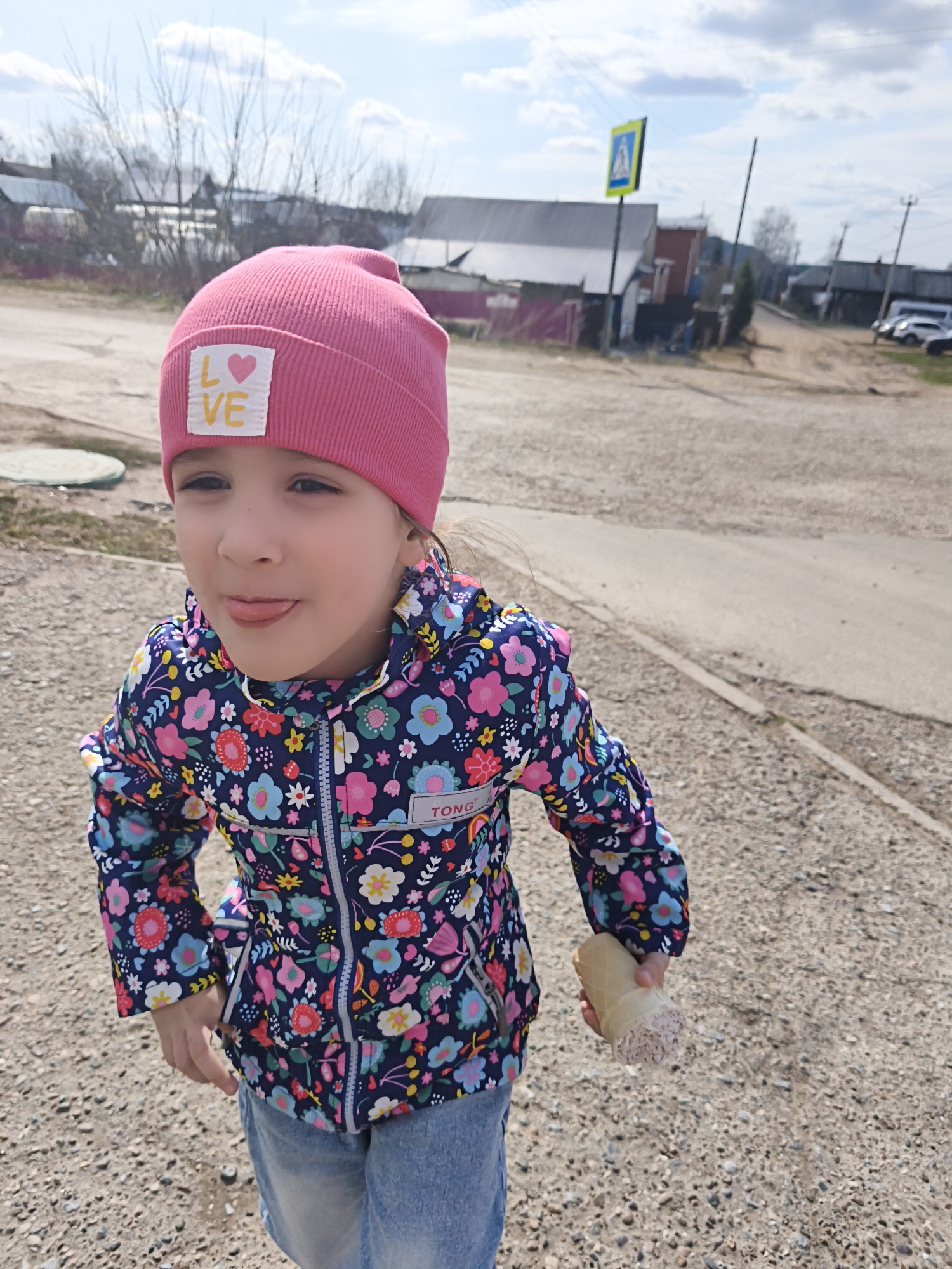 A young girl is captured outdoors, playfully sticking out her tongue. She is looking slightly upward and to the right, with a mischievous expression. She wears a pink knitted beanie with a white label on the front that reads "LOVE" with a small pink heart. Her jacket is dark blue with a vibrant floral pattern of pink, white, blue, and green flowers. A small white label on her jacket sleeve shows the partial text "TONG". She is also wearing light blue denim shorts or jeans. In her left hand, she holds an ice cream cone, which appears to be mostly empty or has some melted contents at the bottom.
The setting appears to be a rural or semi-rural outdoor area on a sunny day with scattered clouds in the sky. The ground beneath her is a mix of dirt and gravel, with a paved road visible behind her. In the background, there are several houses, some with corrugated metal roofs, and bare trees, suggesting either early spring or late autumn. A yellow pedestrian crossing sign with a black symbol of a person walking is visible in the middle distance to the right. Utility poles and lines are also present in the background.