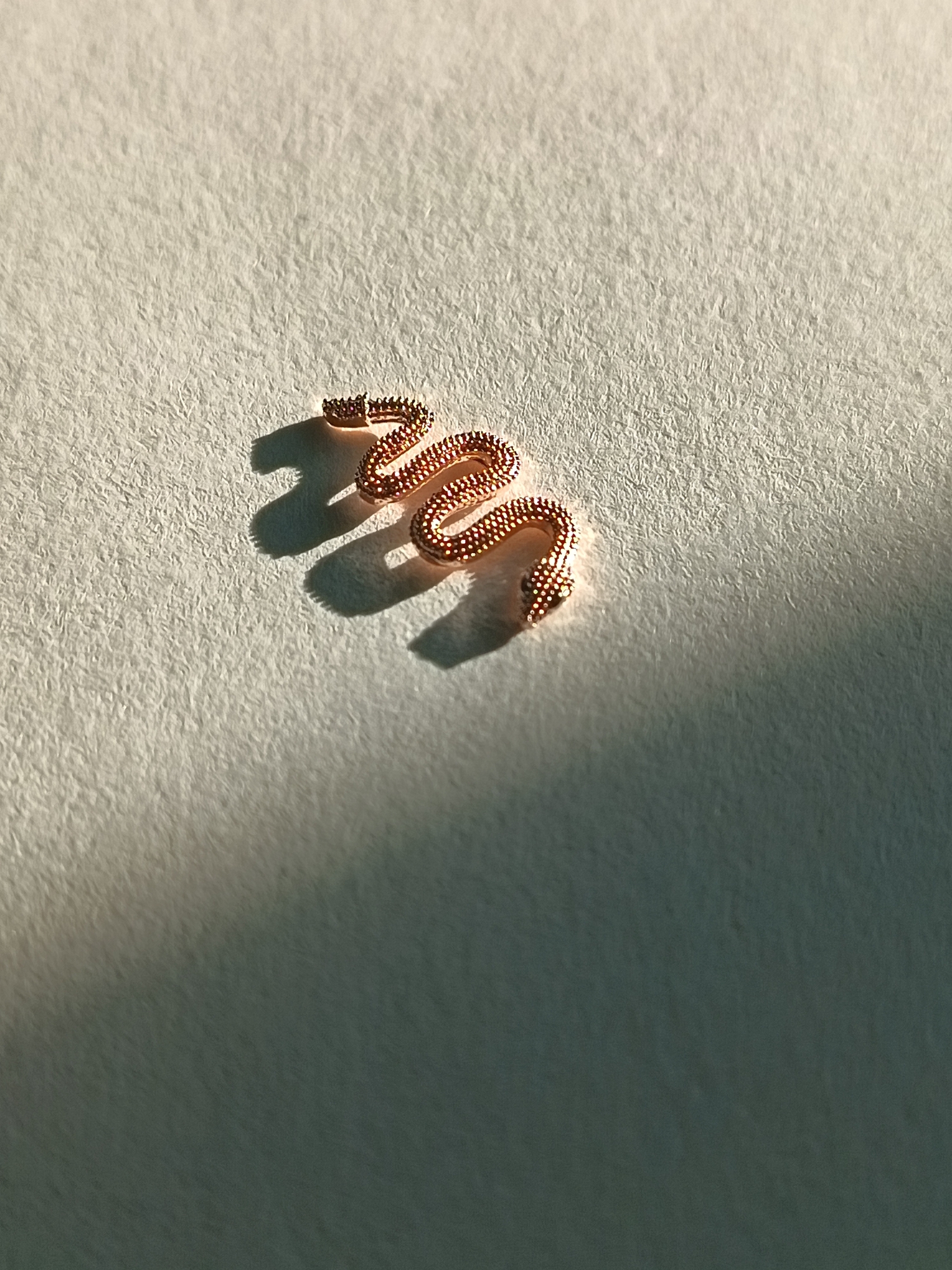 The image displays a close-up, top-down view of a small, coiled, rose gold-colored snake charm or earring. The charm is textured with a beaded pattern and has small, dark accents on its head, likely representing eyes. It is positioned on a textured, off-white surface, possibly paper or fabric, with a noticeable rough grain. Strong, directional lighting casts distinct shadows of the charm across the surface, indicating a light source from the upper left. The overall scene is minimalist and focused on the jewelry piece. The image does not depict any elements that would allow for identification of a specific location in Périgueux, France.