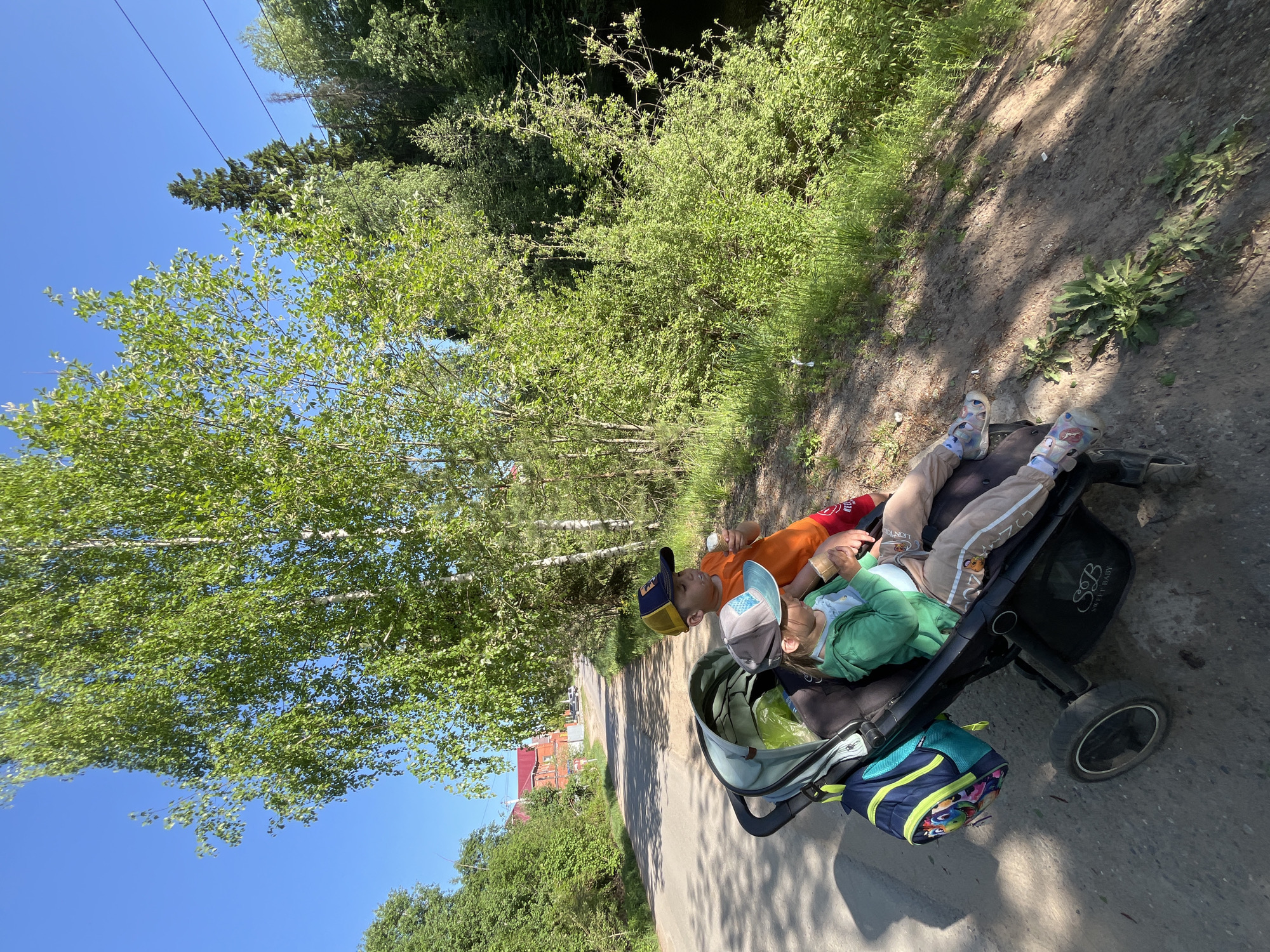 Two children are lying in a stroller on a paved path. The child on top wears an orange shirt and a baseball cap, and the child below wears a green sweatshirt and a baseball cap. A blue and yellow backpack hangs from the stroller. The scene is outdoors, with trees lining the path and a clear blue sky visible above. Power lines are also visible. The location is near the city of Chaikovsky, Russia, as indicated by the "S&B Sweet Baby" logo on the stroller, a brand popular in Russia. A building with a red roof is visible in the background.