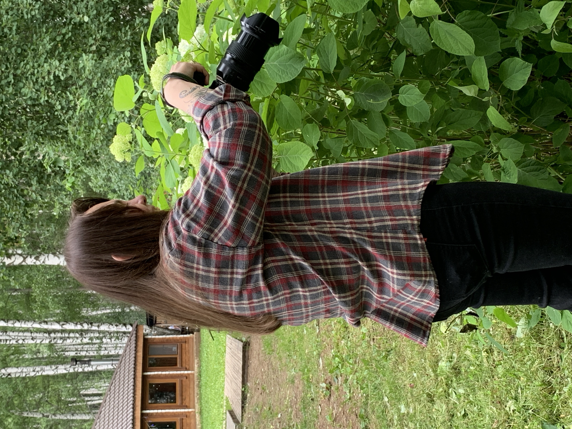 A person with long brown hair is standing outdoors, facing to the right. They are wearing a red and gray plaid shirt and black pants, and are holding a black camera with a zoom lens. The person appears to be taking a photograph of a bush with large green leaves and white flowers. In the background, there is a wooden building with a brown roof and a birch forest. The ground is covered in grass and dirt. The scene is likely in a natural or park-like setting, possibly a dacha or rural area near Chaikovsky, Russia, given the presence of the birch trees and wooden structures.