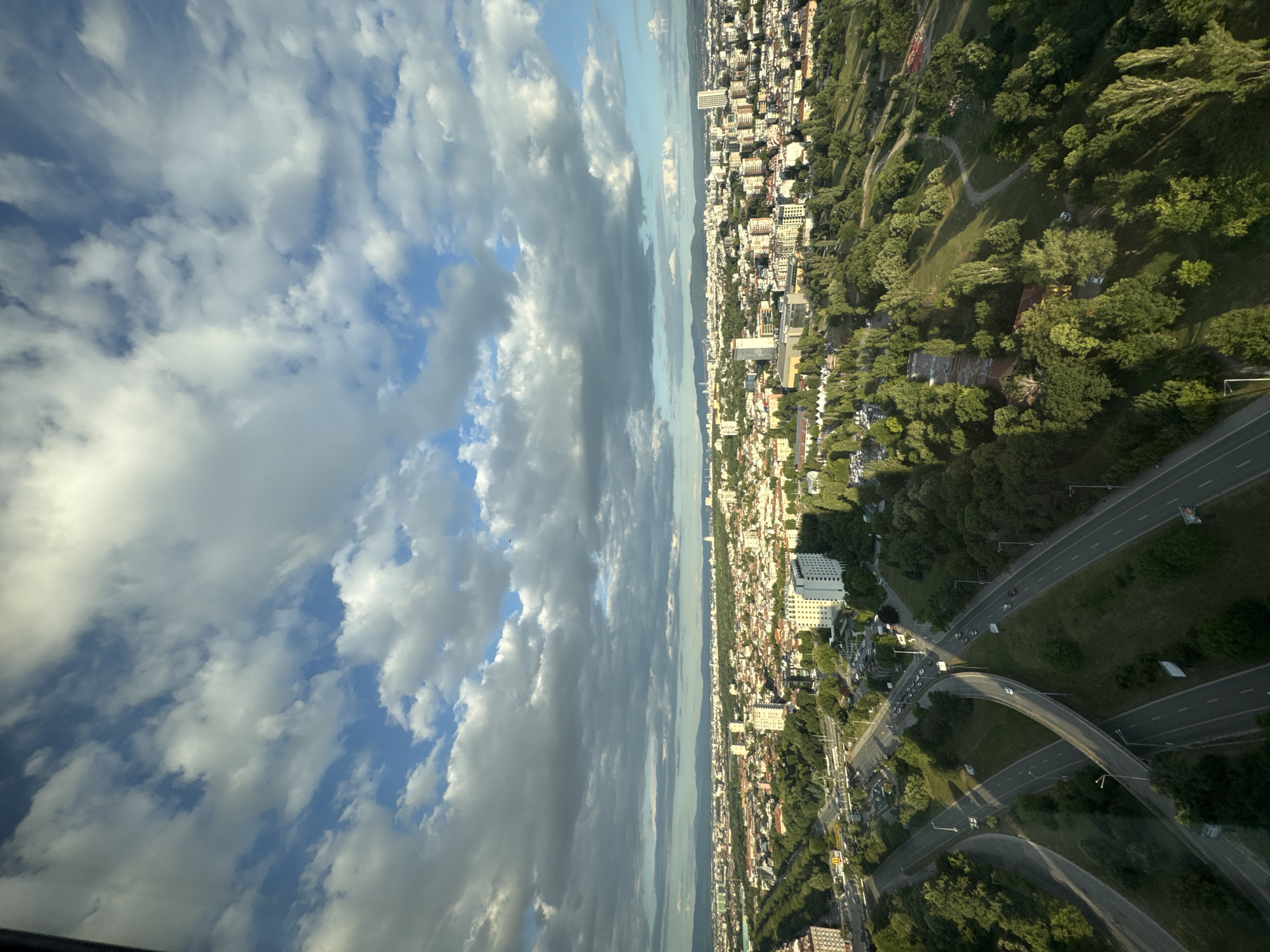 This is an aerial view of a city with a prominent highway interchange in the foreground. The city, identified as Sofia, Bulgaria, features a mix of residential buildings, parks, and some taller structures. The sky is partly cloudy with dramatic formations of white and gray clouds against a blue backdrop. The highway interchange consists of multiple lanes and overpasses, with cars visible on the roadways. Greenery, including trees and grass, is abundant in and around the highway and throughout the urban landscape. The scene appears to be captured during daylight hours, with shadows indicating the sun's position.