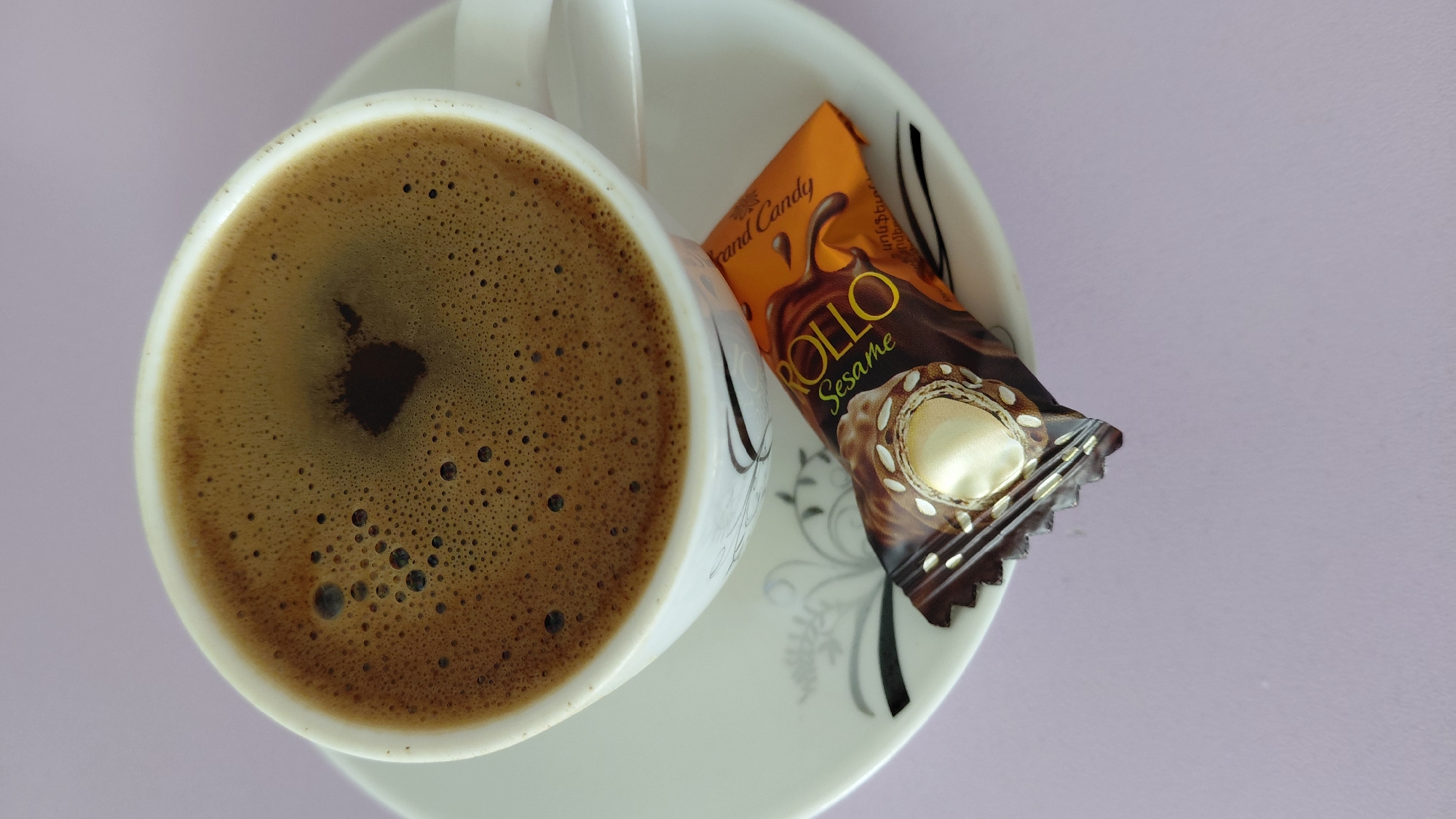 The image shows a white porcelain cup of coffee on a matching saucer. The coffee is dark brown with a layer of foam on top, speckled with small bubbles. To the right of the cup, resting on the saucer, is a single wrapped candy. The candy wrapper is orange and dark brown, depicting a chocolate swirl and the text "ROLLO Sesame" and "Grand Candy". The saucer has a delicate floral pattern in gray and black. The entire arrangement is set against a plain, light purple background. The scene does not contain any people or indicate a specific location within the city of Abovyan, Armenia.