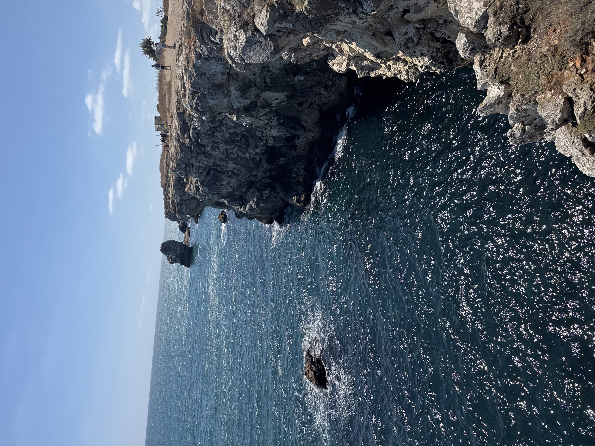 The image depicts a scenic coastal landscape at Tyulenovo, Bulgaria. The foreground is dominated by a steep, rocky cliff face, with rough, weathered rock formations and sparse dry vegetation. Below the cliff, the dark blue sea crashes against the rocks, creating white foam. The water is choppy and reflects the sunlight, creating a sparkling effect.
Further out, the sea stretches towards the horizon under a clear blue sky dotted with a few wispy white clouds. In the distance, several rock formations rise from the water, including a prominent, tall, columnar rock stack.
On the clifftop, a group of people are visible. Some are standing near a small, light-colored building, while others are walking along the edge of the cliff. Two individuals, a male and a female, are seen walking away from the camera on a dirt path. The overall scene conveys a sense of natural beauty and outdoor recreation.