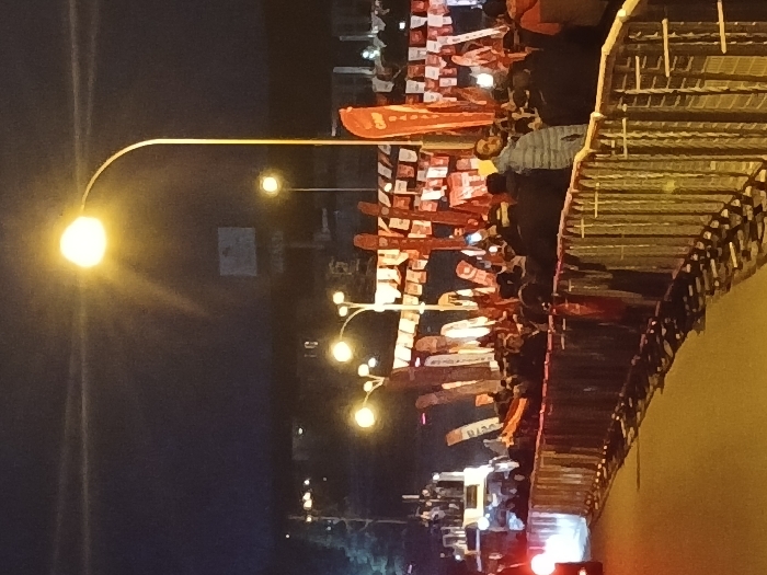 The image shows a crowd of people standing behind a metal barrier on a street in Istanbul, Türkiye at night. The crowd is facing a row of large red and white flags strung across the street. The street is empty in front of the crowd. There are street lights, some with multiple lights, illuminating the scene. There are also vehicles in the distance. The scene is located at the coordinates 41.084, 28.678 in Istanbul, Türkiye.