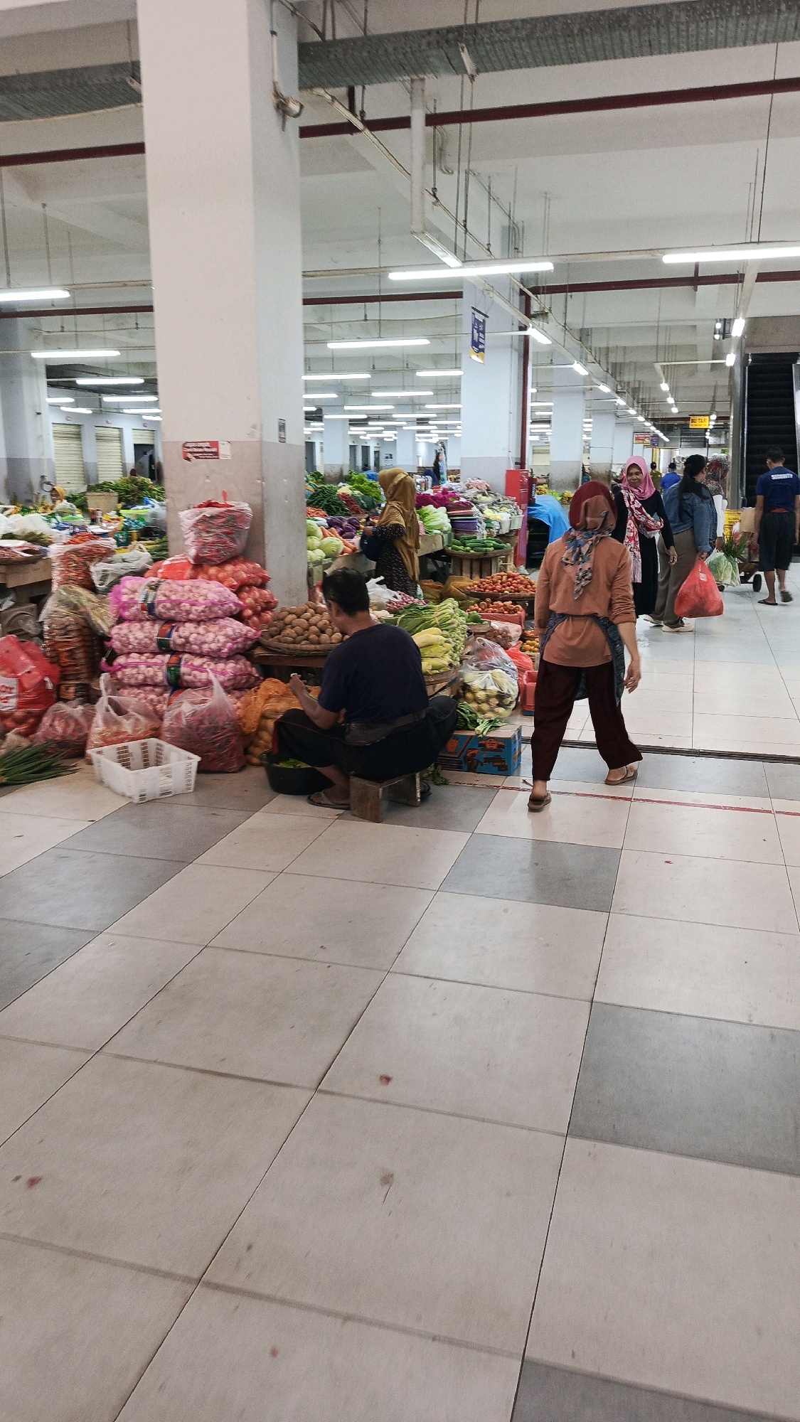 This is a vertical, medium-shot photograph taken inside an indoor market, likely a wet market or produce market. The environment is brightly lit by overhead fluorescent lighting. The floor is tiled in a checkerboard pattern of beige and grey squares. In the foreground, a man wearing a dark blue t-shirt and black pants is seated on a small wooden stool, facing away from the camera and towards a pile of produce. He appears to be sorting or preparing vegetables. To his left are several white plastic baskets and bags filled with produce, including onions and red chilies. Behind him, large stacks of onions are visible. Further into the market, various stalls display a wide assortment of fresh produce, including vegetables and fruits. Several people, predominantly women wearing headscarves, are browsing or working at the stalls. One woman in the mid-ground, wearing a rust-colored top and dark pants, is walking away from the camera. In the background, an escalator is visible, suggesting this may be a multi-level market or part of a larger shopping complex. The overall impression is one of a busy, commercial environment focused on the sale of fresh food. The presence of an escalator and the organized layout suggest it could be a modern market facility.