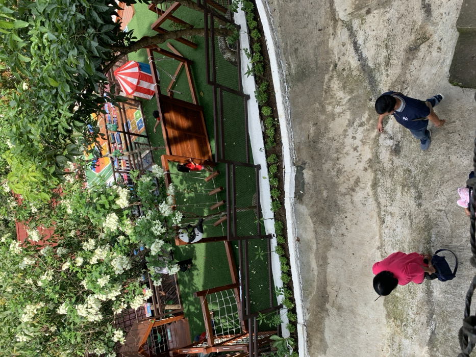 The scene shows a fenced-in playground area in Kecamatan Borobudur, Indonesia. The playground is made of artificial grass with a wooden structure in the background. There is a red and white striped tent in the upper-right corner of the playground. A girl in a red shirt is standing next to a wooden structure in the middle of the playground. Three people are standing on a concrete pathway outside of the playground. A woman in a pink shirt is looking down at the ground and a man in a blue shirt is looking down at the ground. There is a child, obscured by the pathway's edge, wearing a pink and white shirt in the bottom-center of the image. The playground is surrounded by a white concrete curb and a black metal fence, along with a row of green shrubs. There is a large green tree with white flowers in the upper-left corner of the image. This scene is located at a latitude of -7.606 and a longitude of 110.18.