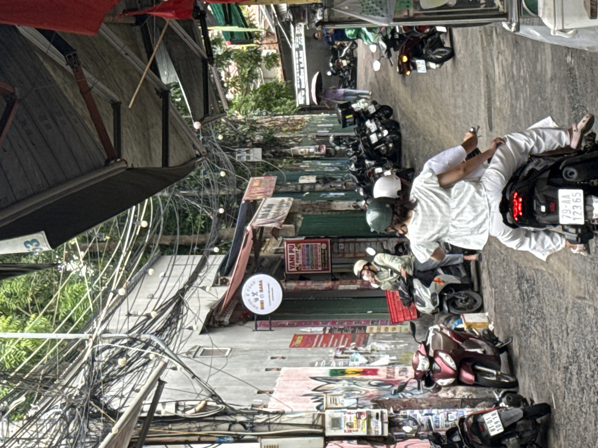 This image depicts a busy street scene in Nha Trang, Vietnam. The foreground features a motorcycle with two passengers, a woman wearing a white top and pants, and a person in the back wearing a green helmet. The motorcycle is black with a white license plate reading "79-AA 123.65". To the left, another motorcycle, red and white, is parked.
The street is paved with asphalt and is lined with buildings on both sides. The buildings have a mix of concrete and metal structures, with many wires and cables crisscrossing overhead, characteristic of urban Vietnamese infrastructure. Several other motorcycles are parked and in motion throughout the scene. Signs in Vietnamese are visible on the buildings, indicating commercial establishments. One sign reads "YANI PU FRUIT".
In the middle ground, a person wearing a helmet and a light-colored shirt is on a white motorcycle, holding a drink. Further down the street, several motorcycles are parked in a row. A person holding a clear umbrella walks in the distance. The overall atmosphere suggests a bustling urban environment with significant motorcycle traffic. The presence of trees and greenery in the background indicates an outdoor setting within the city.