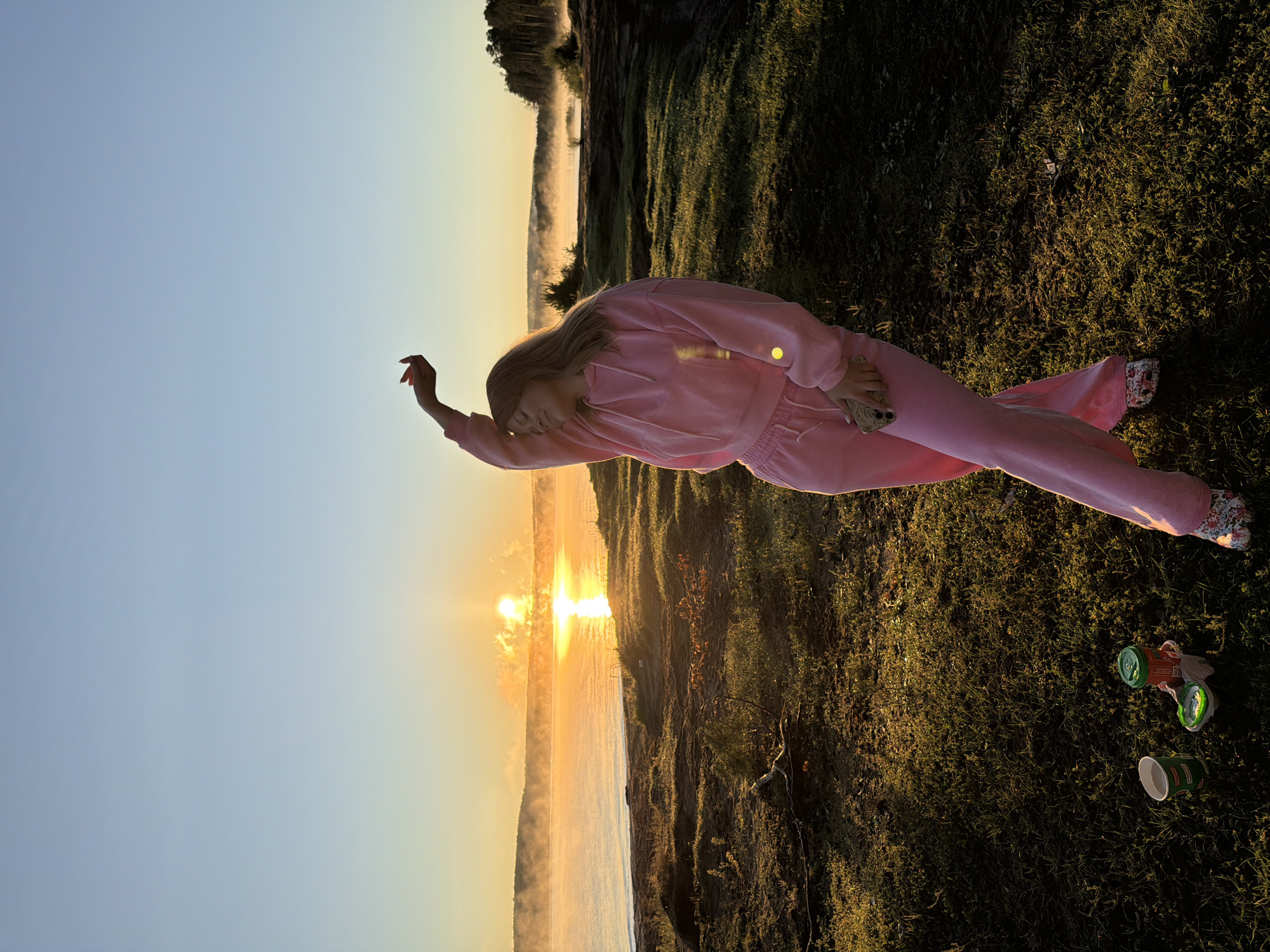 A full-shot, vertical, eye-level, natural light, medium-distance photograph captures a young woman in a pink tracksuit and patterned clogs standing in a grassy area beside a misty body of water during sunrise. The sun, a bright orb, is low in the sky, casting a warm, golden light and reflecting intensely on the water's surface. Mist hovers over the water and the distant tree-lined shore. The woman is positioned in the foreground, facing the right side of the frame with her left arm raised above her head. Her eyes are closed, and her head is tilted back slightly, suggesting a moment of enjoyment or relaxation. Her right hand holds a smartphone. Two disposable cups and a container with a green lid are on the grass in the lower left corner of the frame. The grassy terrain in the foreground appears uneven and sun-dappled. The scene is set along the Kama River in Chaikovsky, Russia, a location known for its picturesque natural landscapes, particularly during sunrise and sunset.