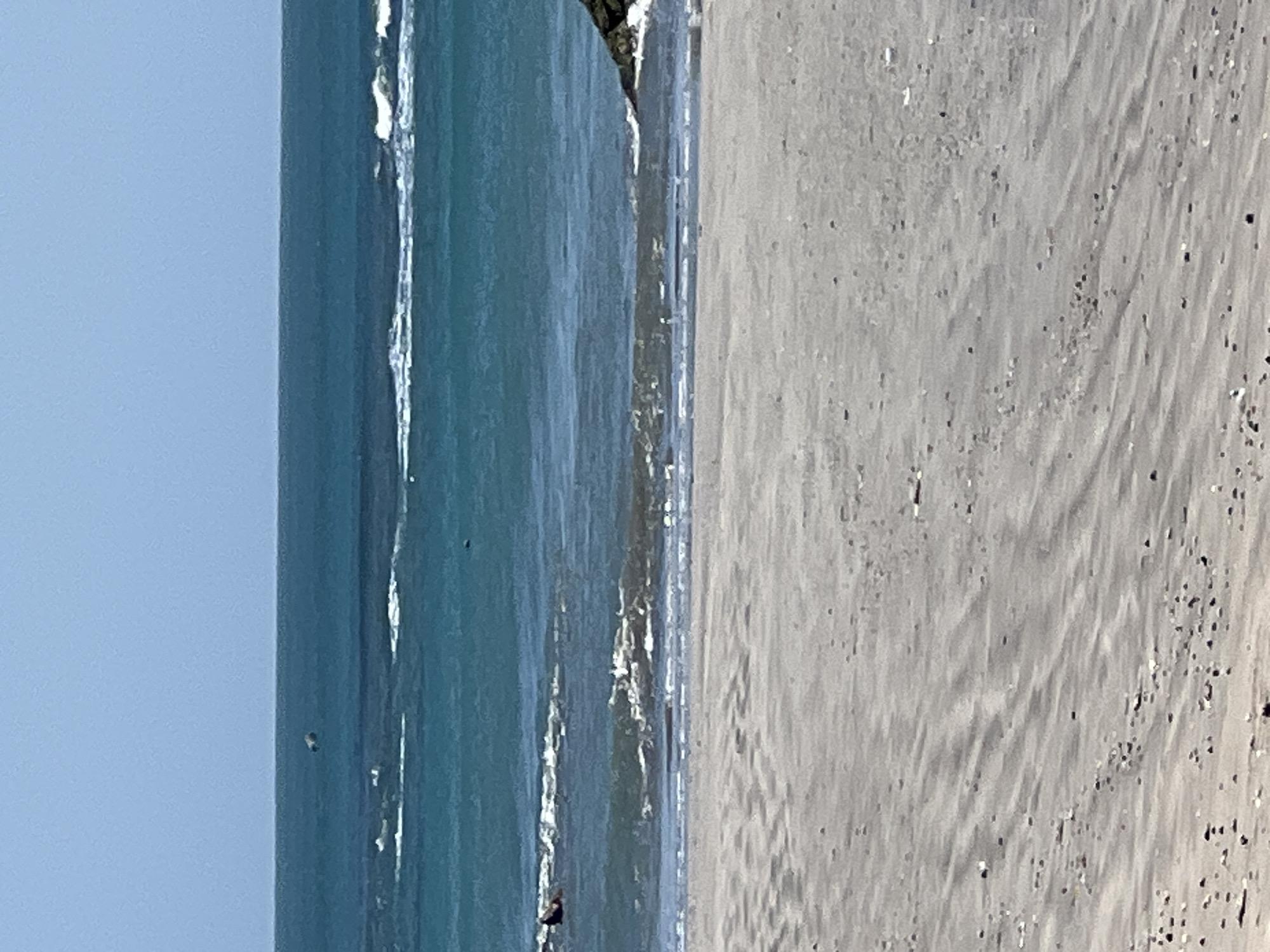 A wide shot of a beach in Kuta, Indonesia, with a person in the water. The sand in the foreground is light gray with subtle ripples from the tide and scattered dark specks. The ocean is a vibrant turquoise, with white waves breaking closer to the shore. In the mid-ground, a person is visible in the water, appearing to be swimming or body surfing. Further out, more white waves are visible, and the horizon is a clear blue sky. A small dark object, possibly a bird or a piece of debris, is seen flying in the distance above the water. On the right side of the frame, a small section of a dark, rocky formation is partially submerged in the water.