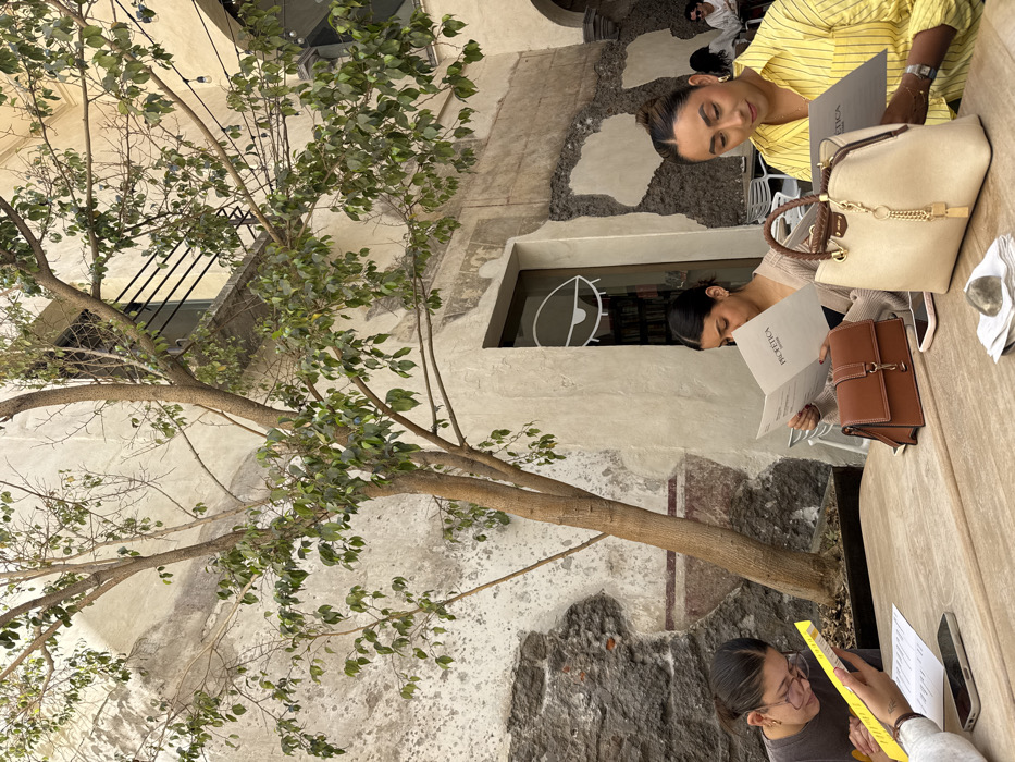 The image displays an outdoor or semi-enclosed patio scene at Profética, Casa de la Lectura, located in the city of Puebla, Mexico. Three women are seated at a light-colored wooden table in the foreground, actively reviewing menus or documents.
On the far left, a woman with dark hair and glasses, wearing a dark top, is partially visible, holding a yellow folder or document. A smartphone is positioned on the table to her right. In the middle-ground, a woman with dark hair pulled back, dressed in a light beige sweater, holds an open menu clearly displaying "PROFETICA." A brown leather handbag, a smartphone, and a folded white napkin are on the table directly in front of her. To the right, another woman with dark hair, wearing a yellow and white striped button-down shirt, is similarly holding an open menu. A cream-colored top-handle handbag with a braided brown handle and gold-colored hardware, a smartphone, and a small, smooth grey stone resting on a white napkin are on the table near her.
A multi-trunked tree with green leaves occupies the center of the frame, its branches extending upwards and across the scene. The background consists of aged, textured walls with areas of peeling light-colored plaster revealing darker stone or brickwork. A distinct doorway or large window behind the central woman features a prominent white stylized eye graphic; bookshelves are visible through this opening. An upper-level window with dark horizontal security bars is partially visible through the tree's foliage. String lights are strung horizontally across the upper right portion of the patio. Blurry figures of additional patrons are discernible at tables in the deeper background.