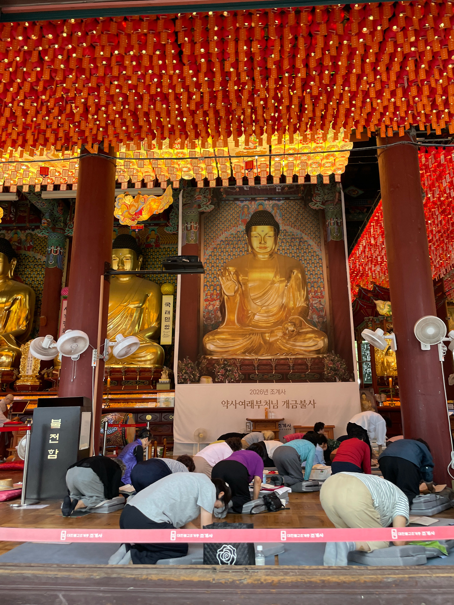 In Seoul, South Korea, inside a temple, a group of people are performing a ritual. The hall is decorated with numerous red and orange lanterns hanging from the ceiling. In the background, there are large, golden statues of Buddha, and a large banner with Korean text. The people are dressed in casual clothing and are kneeling on mats, bowing their heads in prayer or contemplation. Some of them have their foreheads touching the floor. The atmosphere appears to be solemn and spiritual.