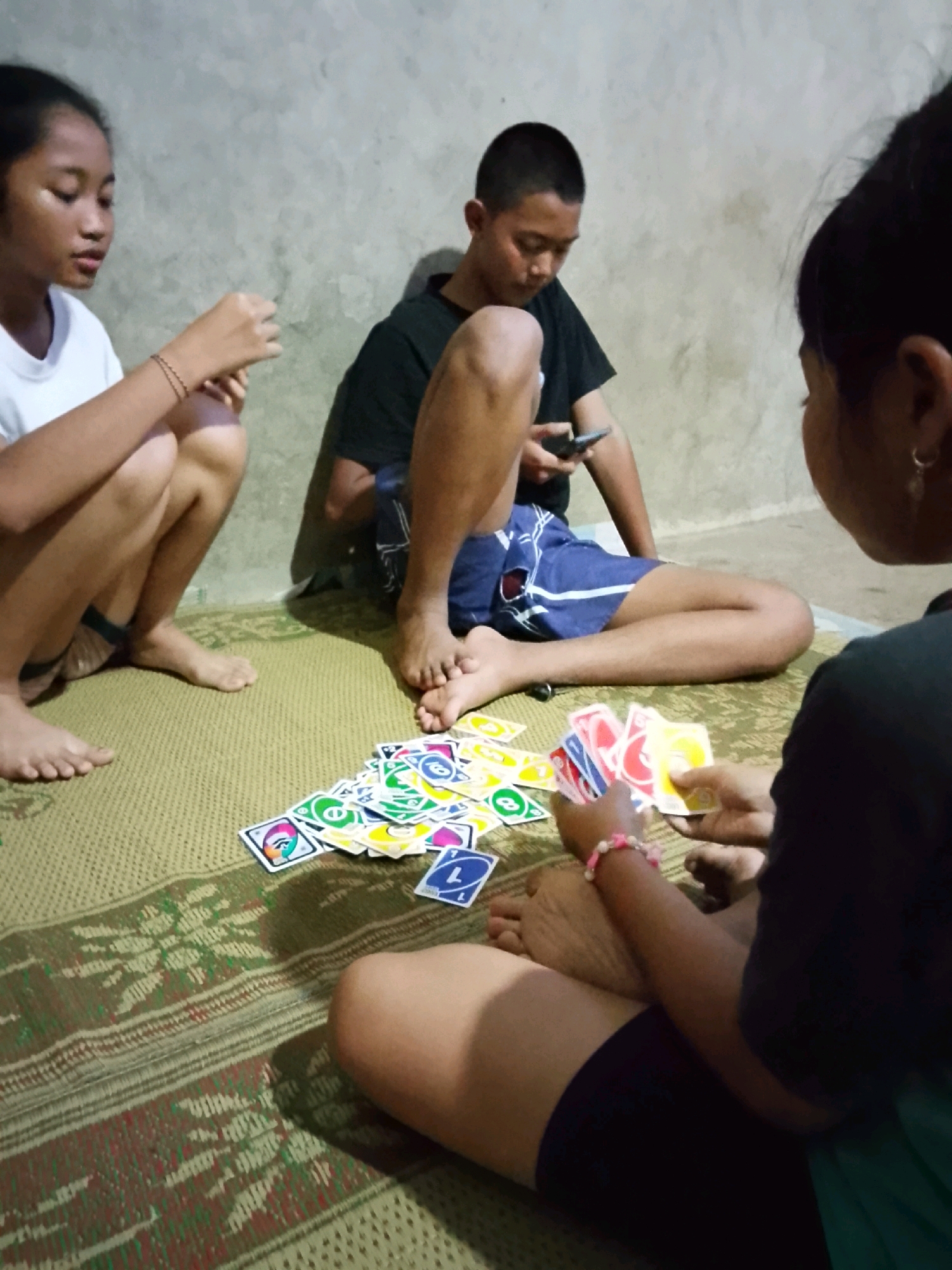 In Gladagsari, Indonesia, three individuals are seated on a woven mat, engrossed in a game of UNO cards. The setting appears to be an indoor space with a concrete wall in the background. A young girl on the left, wearing a white t-shirt, is crouched and holding cards. In the center, a boy in a black t-shirt and blue shorts is seated, looking at a mobile phone. To the right, another person, partially visible from the back, is holding a fan of UNO cards, displaying various colored cards with numbers and symbols. A pile of UNO cards is spread on the mat between the players, suggesting an ongoing game. The lighting is dim, casting shadows on the scene.