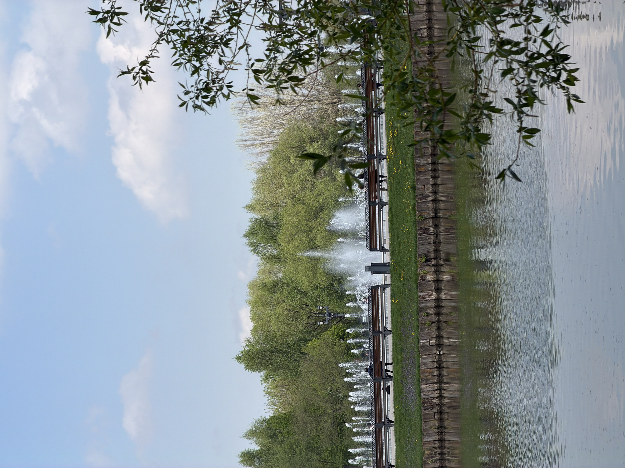 This image depicts a park scene in Moscow, Russia, featuring a pond with a row of fountains along its edge. Benches are placed along the waterside. Trees line the edge of the pond. The sky has scattered clouds. Tree branches are visible along the top of the image, framing the scene. The water reflects the surrounding landscape.