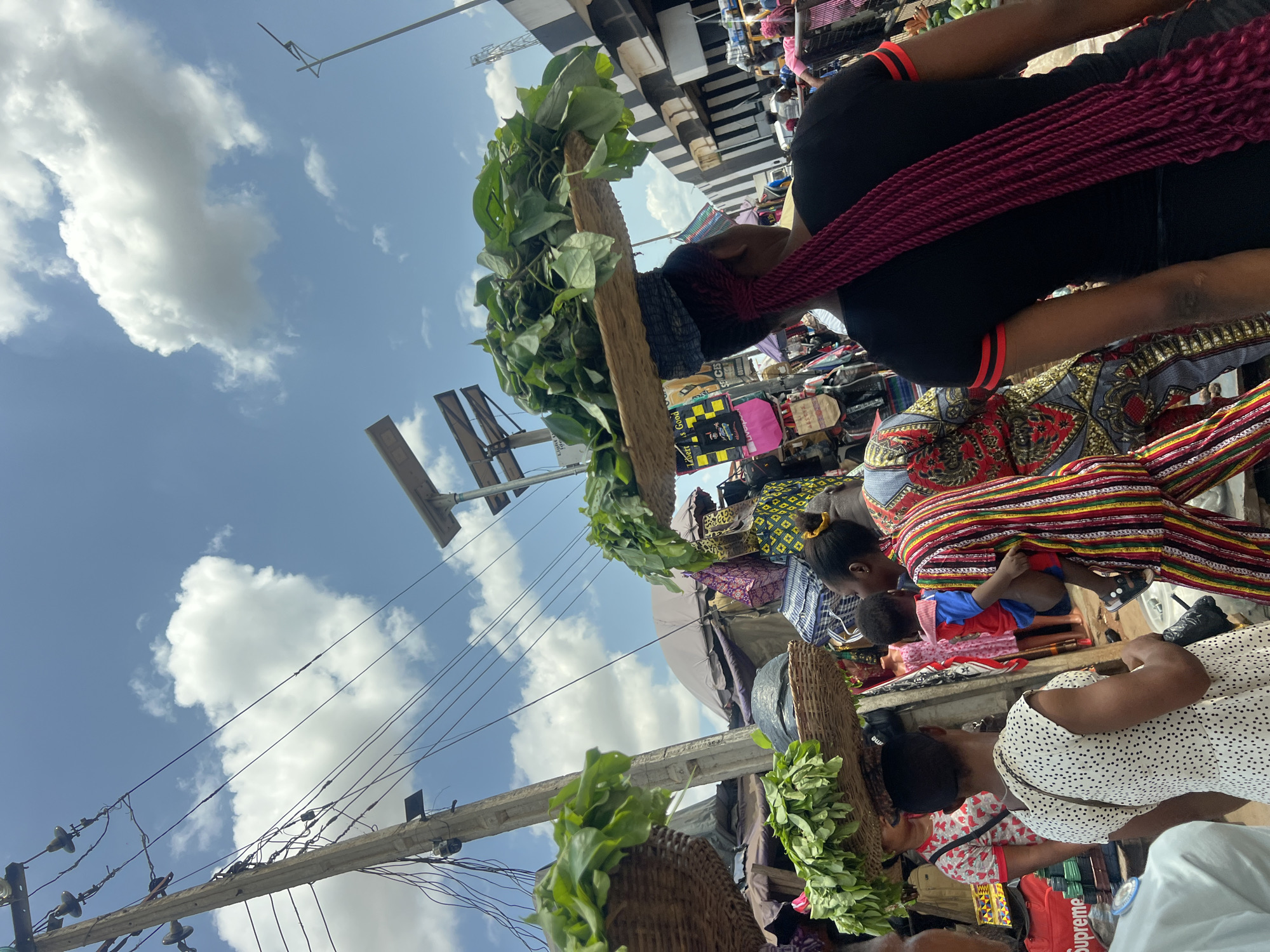This street-level, full-shot depicts a busy outdoor marketplace under a blue sky with scattered clouds. Several individuals are visible in the scene. One woman is wearing a black top with red accents and a colorful patterned skirt. Another woman is wearing a white dress with black polka dots. A child is lying down on a colorful blanket.
The marketplace is filled with various goods and merchandise, including bags and textiles. Baskets filled with green leafy vegetables are prominently displayed.
An overhead structure is visible, with power lines running across the sky. The location is marked as Unknown, Unknown. The scene portrays a typical day at a bustling marketplace.