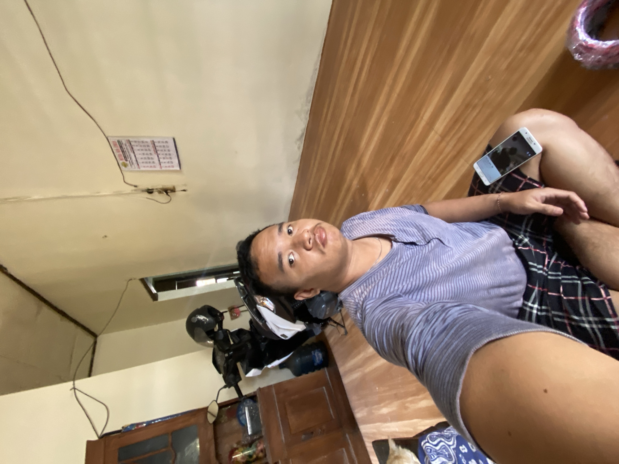 The image shows a man sitting on a wooden floor in what appears to be a room in Sugihwaras, Indonesia. He is taking a selfie, holding his phone in his left hand. He is wearing a light purple striped t-shirt and plaid shorts. Beside him, on his right leg, is a white smartphone. In the background, to the left, is a wooden cabinet with glass doors and a motorcycle with a black helmet on it. A large water dispenser is next to the cabinet. On the wall behind the motorcycle, a calendar is hanging. The walls are painted a light yellow color, and the ceiling is exposed with wooden beams. The floor is made of polished wood planks.