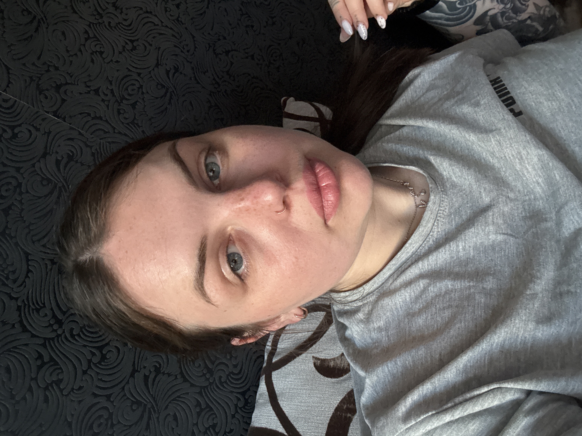 A young woman is captured in a close-up, top-down selfie while lying down in an indoor setting in Chornomorskoe, Ukraine. She has long dark brown hair, blue-grey eyes, and a small hoop nose piercing. Her lips are a light pink, and freckles are visible on her nose and cheeks. She wears a light grey t-shirt with a visible, reversed "PUMA" logo across the chest, suggesting a front-facing camera or mirror shot. A delicate silver chain necklace adorns her neck. Her fingernails are long, almond-shaped, and adorned with a light, decorative pattern, possibly white marble or glitter. A dark, intricate tattoo sleeve is visible on her right arm/shoulder area. She gazes directly at the camera with a neutral expression. The background consists of a dark grey or black patterned fabric, likely a bedspread or sofa, featuring swirling, ornate designs. Another section of fabric with a lighter grey background and brown geometric patterns is also partially visible, possibly part of a cushion. The overall impression is one of a relaxed, informal moment.