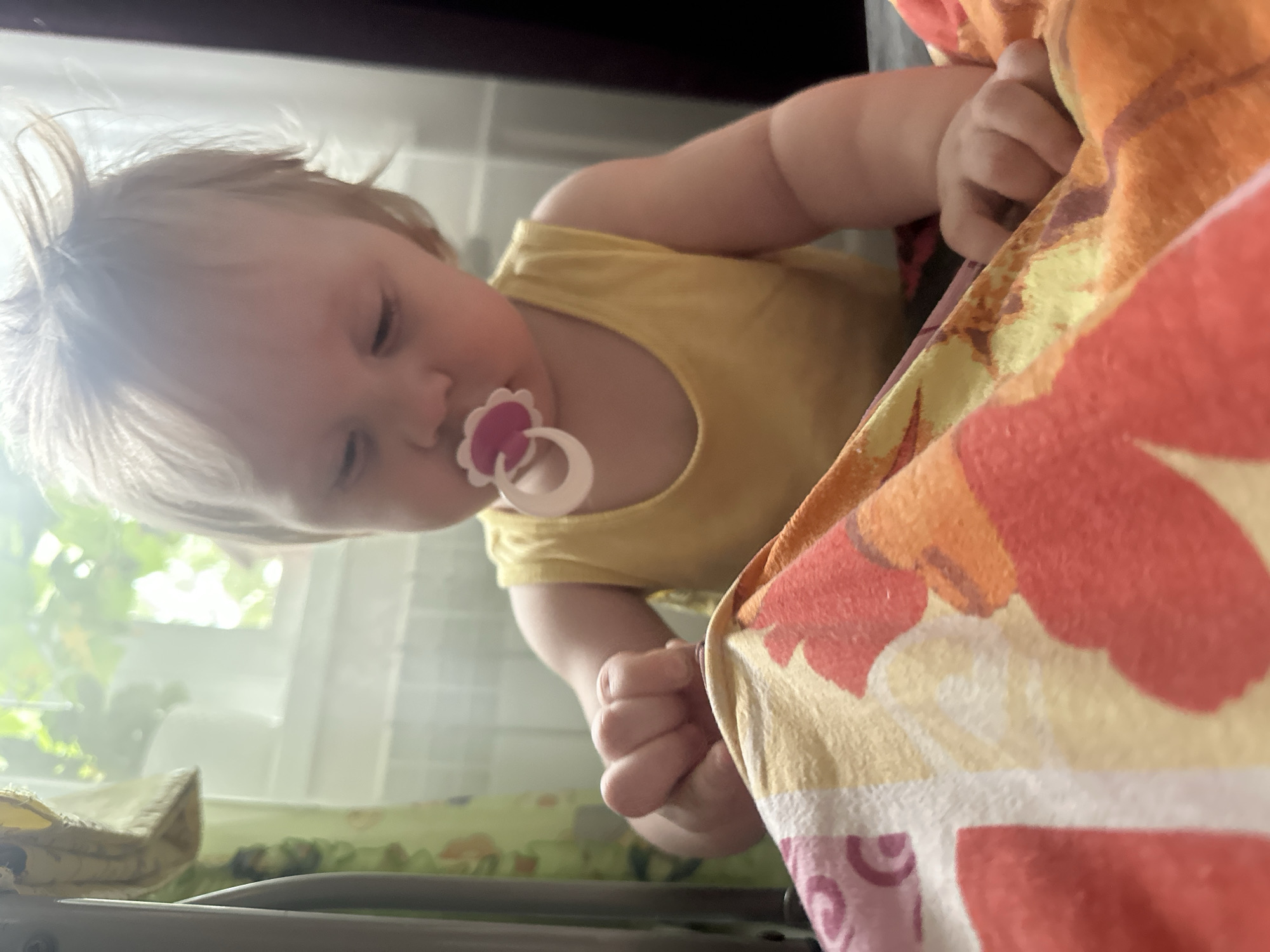 A baby wearing a yellow tank top and a pacifier is sitting on a colorful blanket with a floral pattern. The baby's hands are on the blanket, and the baby is looking down at the blanket. The background is blurred, but it appears to be a room with a window. The image appears to be taken indoors. The location is the city of Sayanogorsk, Russia.