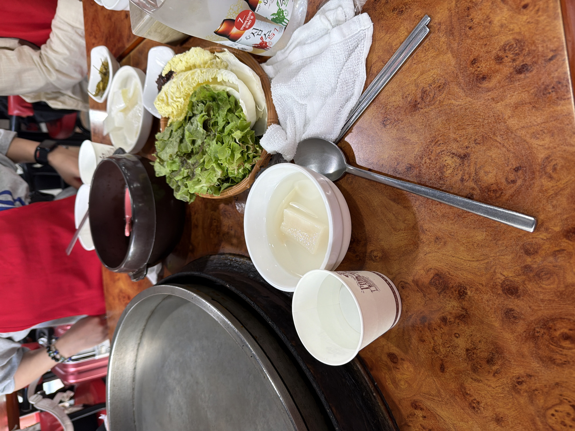 The image depicts a close-up, overhead view of a table setting at a restaurant, likely in Chuncheon-si, South Korea. A person, partially visible at the top, wears a red apron and a grey shirt, with beaded bracelets on their left wrist. The table surface is a warm, polished wood with a distinctive grain pattern.
The primary focus is on a meal laid out on the table. In the foreground, to the left, is a white paper cup filled with clear liquid, possibly water. Next to it, to the right, is a white bowl containing a light-colored broth with rectangular pieces of white food, likely radish or tofu. A silver spoon rests on the table beside the bowl. Further to the right, a basket holds fresh green lettuce and pale yellow napa cabbage leaves, arranged for wrapping food. A small white dish contains a dark red paste, likely a dipping sauce. Two metal chopsticks lie parallel to each other on a white cloth. Behind these items, a dark brown ceramic bowl is visible, containing a pinkish substance. To the far right, a clear plastic bottle with Korean labeling, featuring images of beer bottles, is present. In the background, a large, circular metal pan, likely for cooking, is set on a black stand. Another person's hands are visible behind the pan, suggesting they are preparing or serving the food. The overall impression is of a meal in progress, possibly a Korean BBQ or hot pot dish.