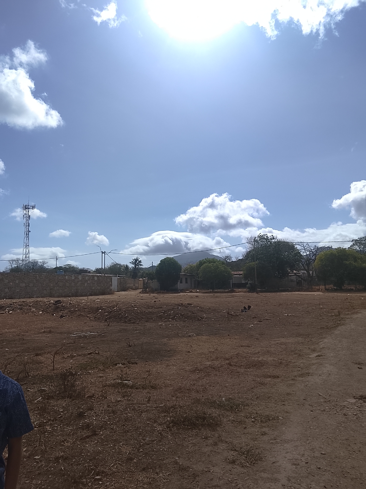 The image shows a rural scene in Altagracia, Venezuela, under a bright, partially cloudy sky with a visible sun glare. In the background, a low mountain is partially obscured by a large cloud. Several houses and trees are scattered throughout the midground. A stone wall and a communications tower are visible on the left side of the frame. The foreground is dominated by a dry, barren field with sparse vegetation and a dirt path. A person wearing a blue shirt is partially visible on the lower left side of the image.
