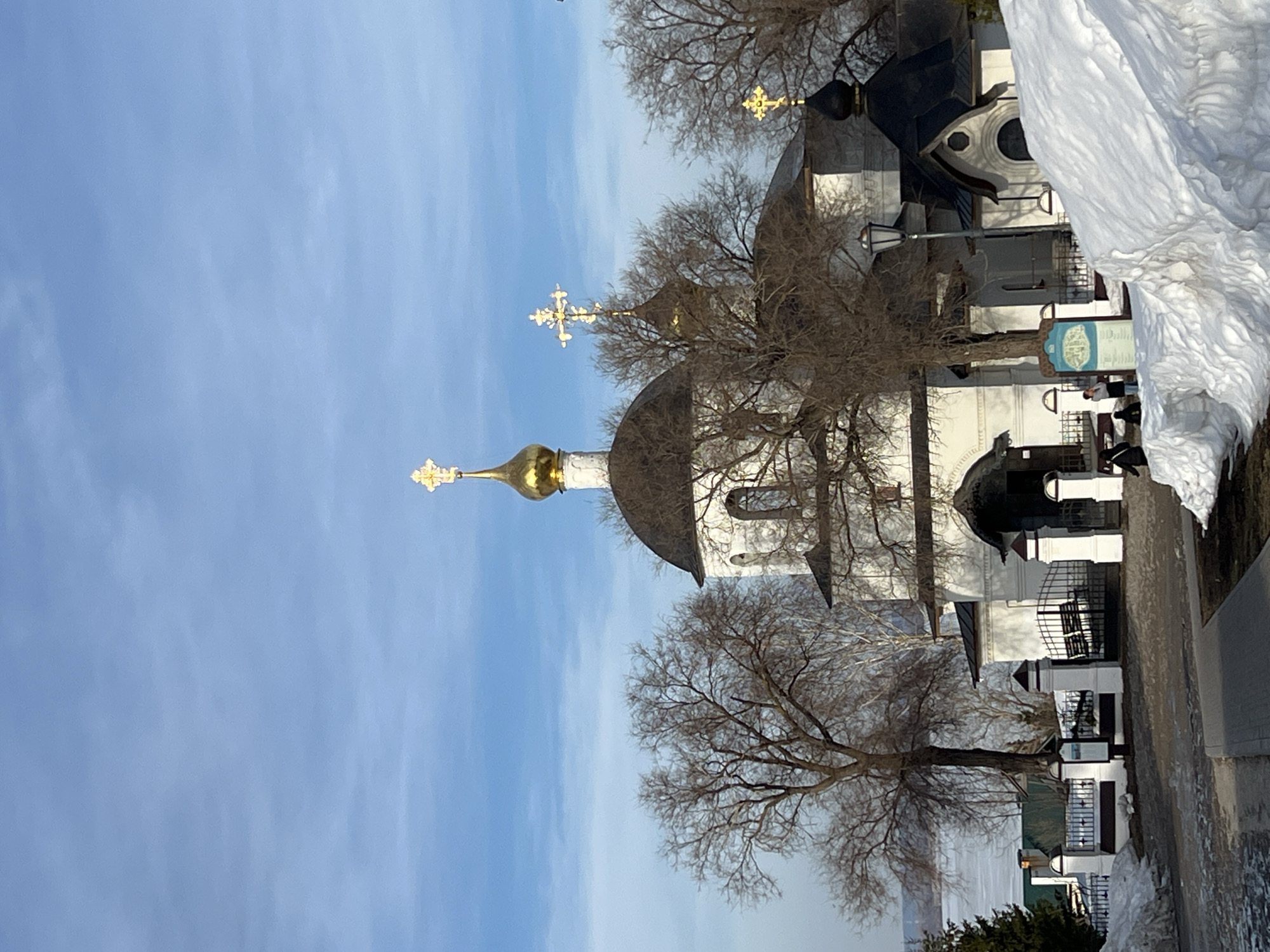 The image displays a winter scene of the Sviyazhsk Assumption Monastery complex located on the island town of Sviyazhsk, Russia. The central focus is a group of Russian Orthodox structures, predominantly featuring white walls and dark, sloped roofs. A prominent, large golden onion dome, topped with a golden Orthodox cross, stands centrally. Additional smaller golden and dark onion domes, also crowned with golden crosses, are visible across the complex. The architectural style includes arched windows and entranceways.
In the foreground, a cleared paved pathway leads towards a black wrought iron gate, supported by white stone pillars. Large, uneven snowdrifts are accumulated on the right side of the foreground, creating a significant white mass. Bare deciduous trees, characteristic of winter, are distributed around and behind the monastery buildings.
Several individuals are present in the mid-ground. Three figures are observable near a blue information signpost positioned beside the path on the right. Other indistinct figures are partially visible to the left, near the entrance path. A black streetlamp with a decorative lantern head is situated on the right side, between a snowdrift and a smaller monastery building which features a dark onion dome.
In the distant background, a wide, frozen body of water is visible beneath a bright, light blue sky with scattered wispy clouds, indicating clear winter weather.