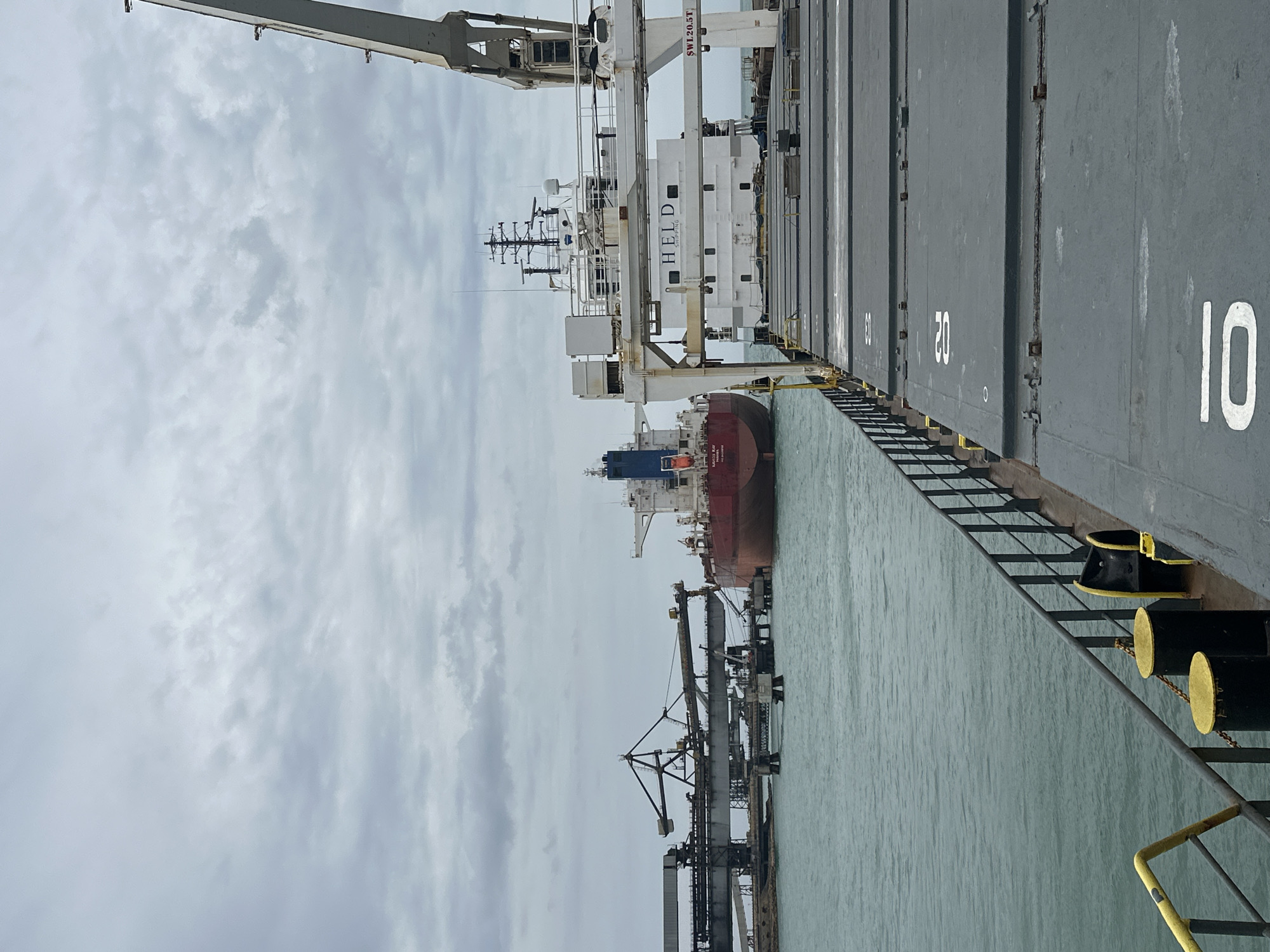 This forensic image captures a maritime industrial scene in Uribia, Colombia, under a uniformly overcast sky. The viewpoint is from the deck of a large cargo vessel, looking forward and towards the port side. The foreground displays the gray, textured deck with several large hatch covers featuring prominent white numerical markings, specifically "10" and "20". A dark metal railing with a yellow top runs along the left edge of the deck, terminating near black cylindrical bollards topped with yellow caps. The water adjacent to the vessel is a murky teal color.
In the midground, to the right, a white cargo ship, identified by the name "HELD SHIPPING" on its superstructure, is visible. This vessel features a complex array of antennas and radar equipment on its mast and a white crane arm marked with "SWL 20.5T". Towards the center-left midground, a large red-hulled bulk carrier, displaying "NAVIOS RAY" and "PANAMA" on its stern, is positioned in the water, appearing to be either anchored or navigating. Adjacent to this vessel on the far left, a substantial industrial bulk loading facility extends over the water, characterized by multiple interconnected metal conveyor structures supported by concrete pylons. No human figures are discernible within the frame.