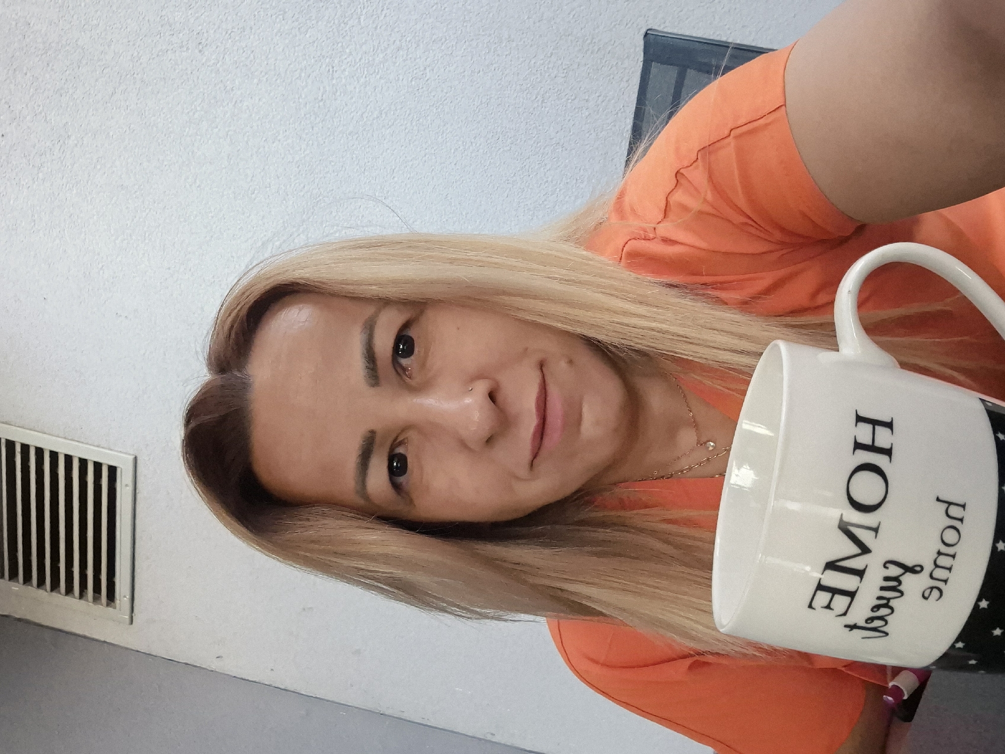 A medium shot image shows a woman with blonde hair and an orange shirt sitting at a table, holding a white mug that says "HOME sweet HOME" in black lettering. She is looking directly at the camera with a slight smile. In the background, there is a white wall with a vent and a chair. The lighting is natural and appears to be from an outdoor setting.