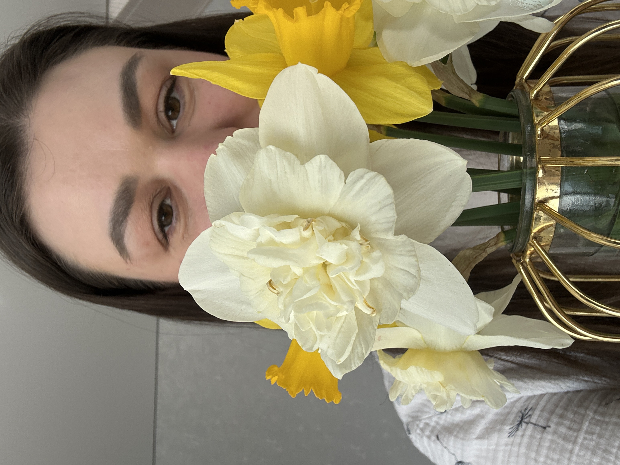 A woman with dark hair and eyebrows is partially visible behind a vase of white and yellow daffodils. The vase is gold and has a geometric design. The woman's eyes are brown. She is wearing a white shirt with a small, dark pattern. The background is a light gray wall. The location in the city of Chaikovsky, Russia, cannot be determined from the image.