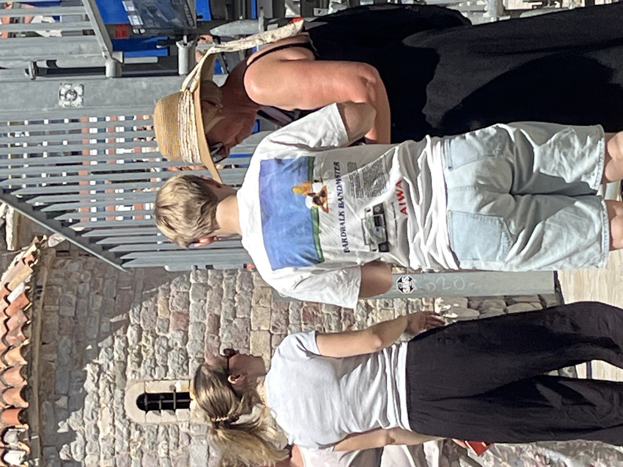 The image captures three individuals on a sunlit street in Budva, Montenegro, with a stone building and metal fencing in the background. A woman with blonde hair pulled into a ponytail, wearing sunglasses and a light-colored t-shirt with black wide-leg pants, stands on the left. She is holding a red object, possibly a phone or wallet, in her right hand. In the center, a person with short blonde hair, wearing a white t-shirt with a graphic print and light blue denim shorts, is facing away from the camera. The graphic on the t-shirt appears to be a beach scene with text that includes "BOARDWALK BANDMASTER" and "AIWA." To the right, an older woman wearing a wide-brimmed straw hat and sunglasses, a black tank top, and a black dress or skirt is interacting with the person in the center. The environment features a textured stone wall with a small arched window and a corrugated metal fence structure. The lighting suggests it is daytime, with strong shadows indicating a bright sun.