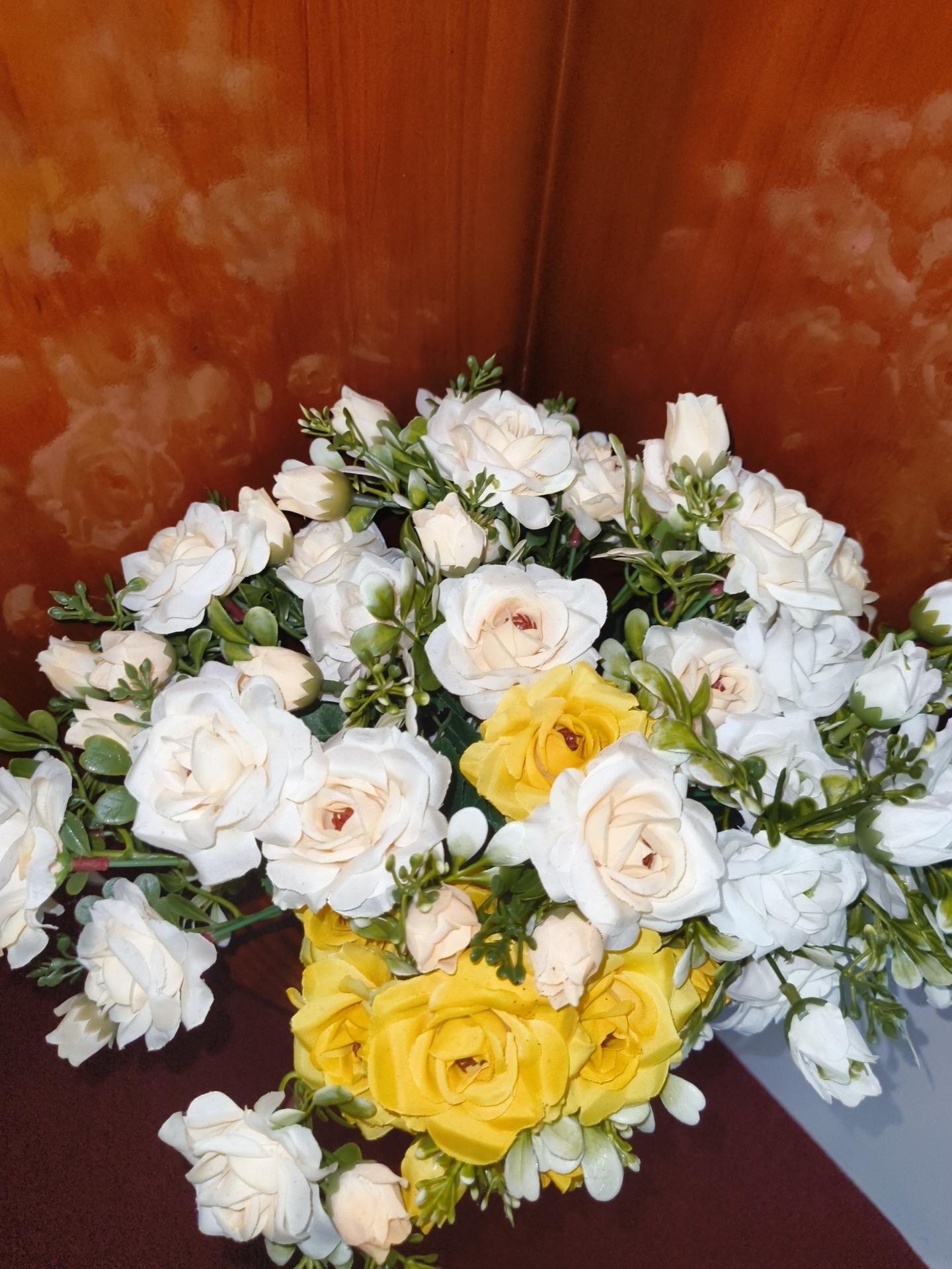 A close-up, high-angle, medium shot captures a bouquet of artificial flowers, primarily white and yellow roses, arranged in a vase. The flowers are a mix of open blooms and buds, interspersed with artificial green leaves and stems. The bouquet is positioned against a backdrop of a dark brown, glossy surface, likely a wooden cabinet or wall, which exhibits subtle lighter brown swirling patterns, suggesting a wood grain texture or a decorative finish. The lighting is artificial and seems to be from above and to the right, casting some shadows and highlighting the textures of the flowers and the background. No people are visible in the image, and no specific landmarks or environmental cues indicate a location in Phu Thuy Ward, Vietnam. The scene is static and presents a still life arrangement.