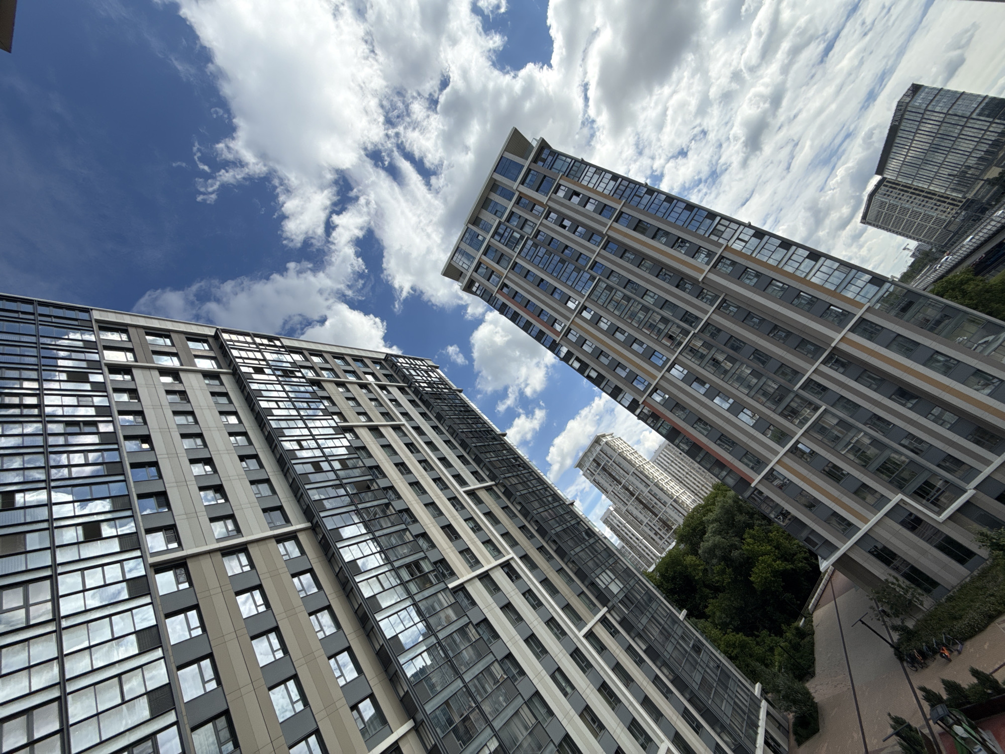 The image captures a view of modern high-rise residential buildings against a partly cloudy blue sky, likely in Moscow, Russia. The perspective is looking upwards from a low angle, emphasizing the scale and architecture of the buildings. The building in the foreground on the left is a tall, multi-story structure with a facade of glass windows and light-colored panels. The building on the right is also a high-rise, characterized by its vertical stripes of dark and lighter colored panels and numerous windows. In the background, several other apartment blocks are visible, some appearing lighter and more distant. The presence of trees suggests some greenery within the urban landscape. The sky is a vibrant blue with scattered white clouds, indicating fair weather. No people are visible in the image. The scene is static, depicting a typical urban residential environment.