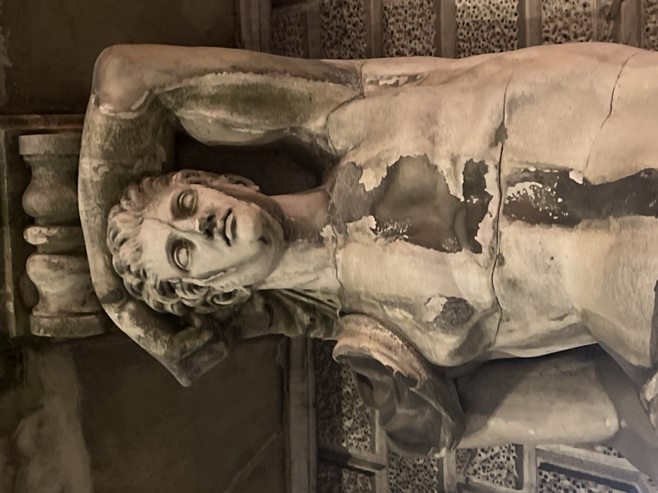 A close-up shot shows a decaying classical marble statue of a male figure, likely a caryatid, lying on its side. The figure's head, torso, and upper arms are visible, resting on an ornate pillar. Damage is evident, with significant surface peeling and cracks. The statue's head features curly hair and distinct facial features. The background includes elements of decorative stonework.