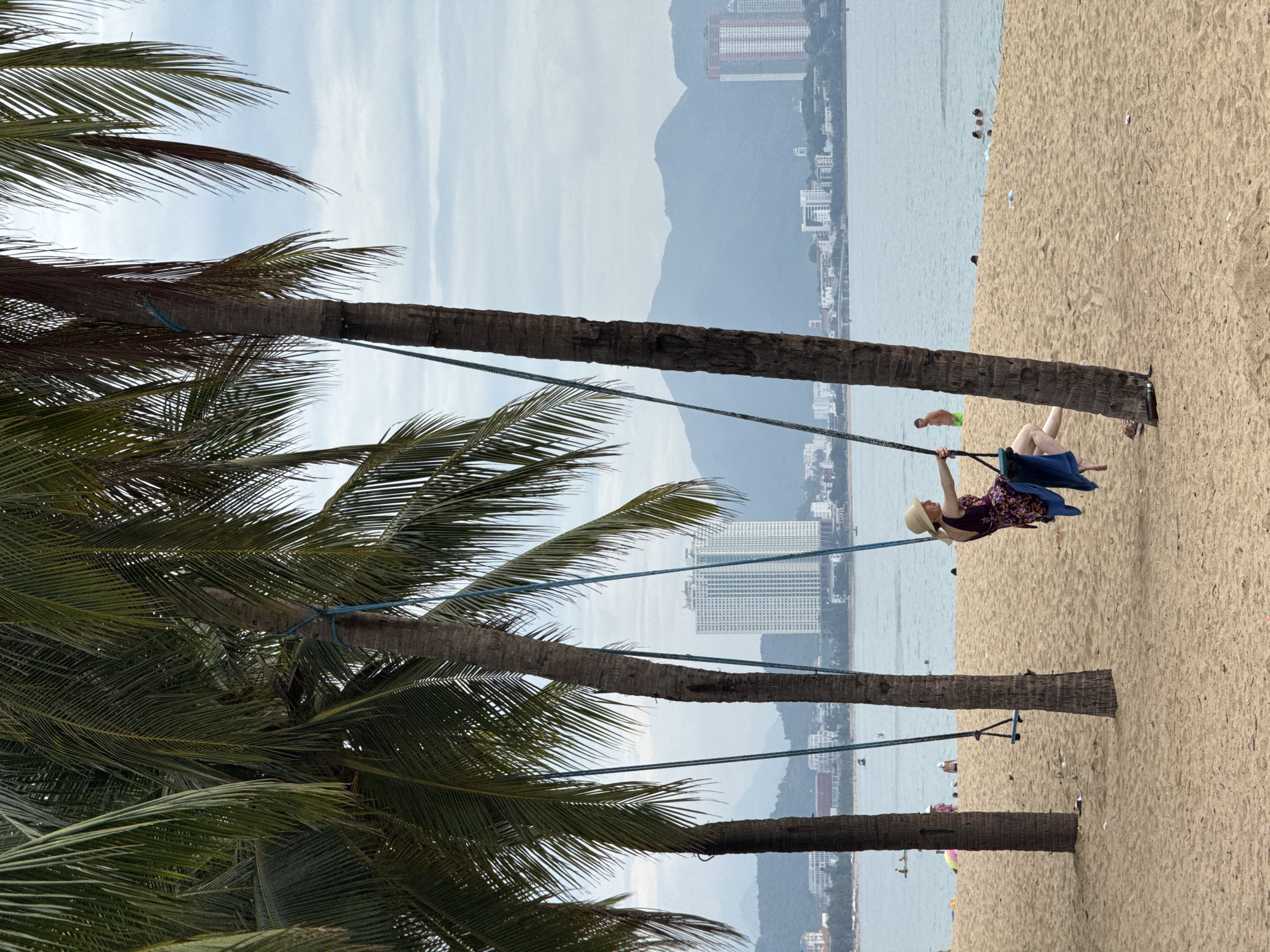 In Nha Trang, Vietnam, a woman in a straw hat and a floral dress is swinging on a rope swing suspended between two palm trees on a sandy beach. She is facing left, and her legs are bent. A blue towel is draped over her lap. The beach is populated with a few other people swimming in the ocean or relaxing on the shore. In the background, a city skyline with tall buildings and mountains can be seen under a cloudy sky. The sea is calm and appears to be a light blue color.