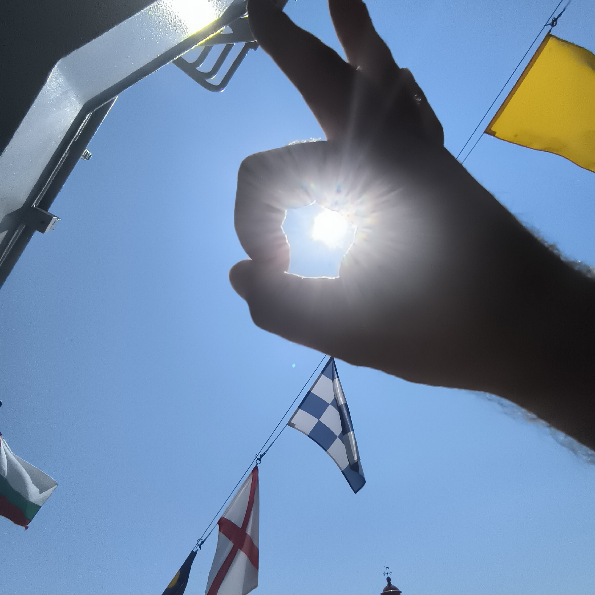 The image, captured on a bright, sunny day in Varna, Bulgaria, shows a hand forming an "okay" symbol with its index finger and thumb, silhouetted against the sun. The sun's rays are visible, creating a starburst effect as they pass through the gap formed by the fingers. Below the hand, a string of nautical flags is suspended against a clear blue sky. These flags include a yellow flag, a checkered blue and white flag, and a white flag with a red cross. In the distance, the dome of a building, possibly a church or a historical structure, is partially visible. The overall scene suggests a festive or maritime atmosphere, possibly near the Varna port or along the seaside promenade.