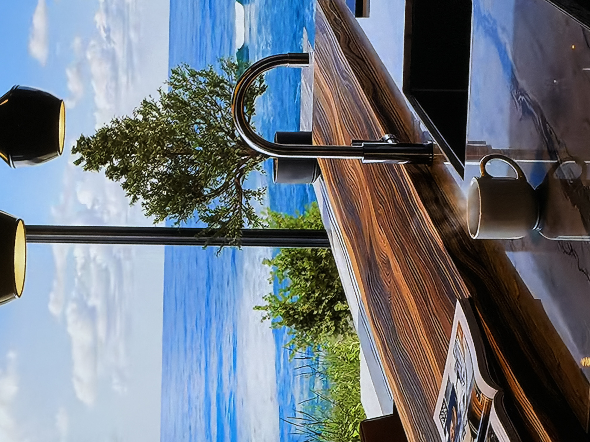 This image, oriented horizontally, presents an indoor scene with a panoramic outdoor view. A clear blue sky with scattered white clouds stretches above a vast expanse of blue water, likely a sea or large lake. Lush green trees and bushes fringe the water's edge, enhancing the natural vista.
The foreground showcases a stylish interior featuring a warm-toned wooden counter or bar with a distinct wood grain pattern. A modern, polished chrome gooseneck faucet is centrally positioned on this surface. To the right, a simple white ceramic mug rests on a dark, reflective countertop, its reflection clearly visible. Two black pendant light fixtures, emitting a soft yellowish glow, are partially visible in the upper left corner. An open book or magazine with undecipherable text lies on the wooden counter, suggesting recent activity or a moment of leisure.
The scene is bathed in bright daylight, indicating clear weather. Although no people are present, the setting evokes a sense of tranquility and offers a serene space for relaxation or contemplation, connecting the modern interior with the expansive natural beauty outside. The provided location context is Sofiivska Borshchahivka, Ukraine, though the visual suggests a prominent coastal or lakeside setting.