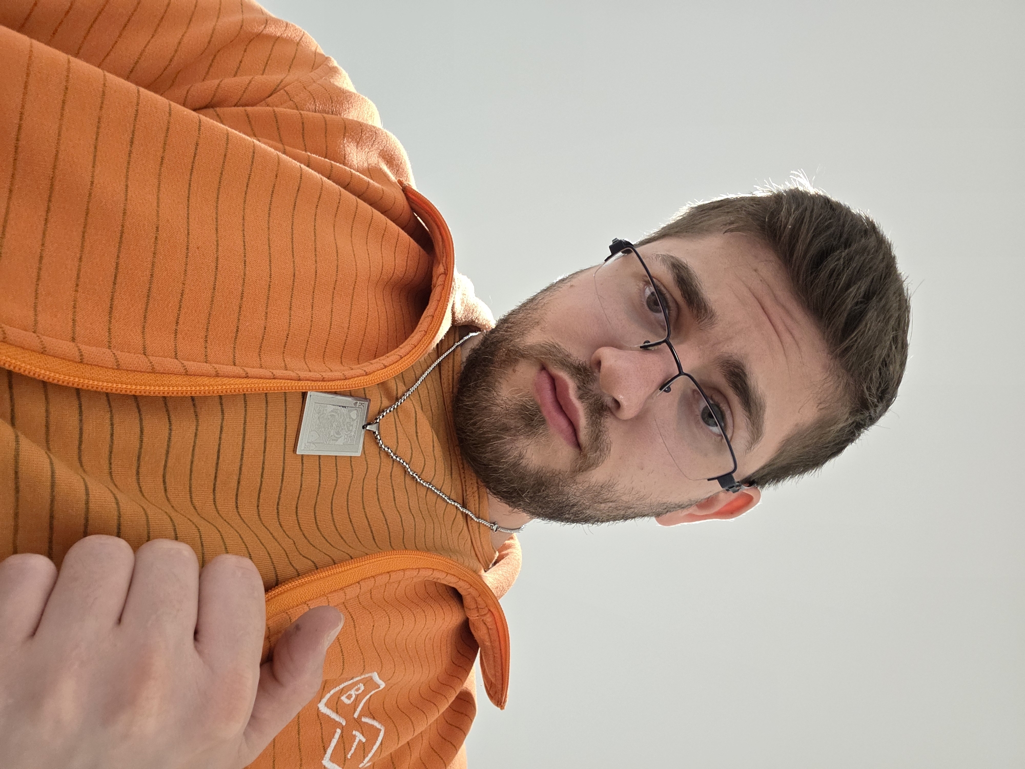 A man with brown hair and a beard, wearing glasses and an orange striped hoodie, looks down at the camera. He is wearing a silver necklace with a rectangular pendant. His hand is visible in the lower left corner, touching the hoodie. The background is a clear, light gray sky. The image does not appear to be in Plovdiv, Bulgaria.