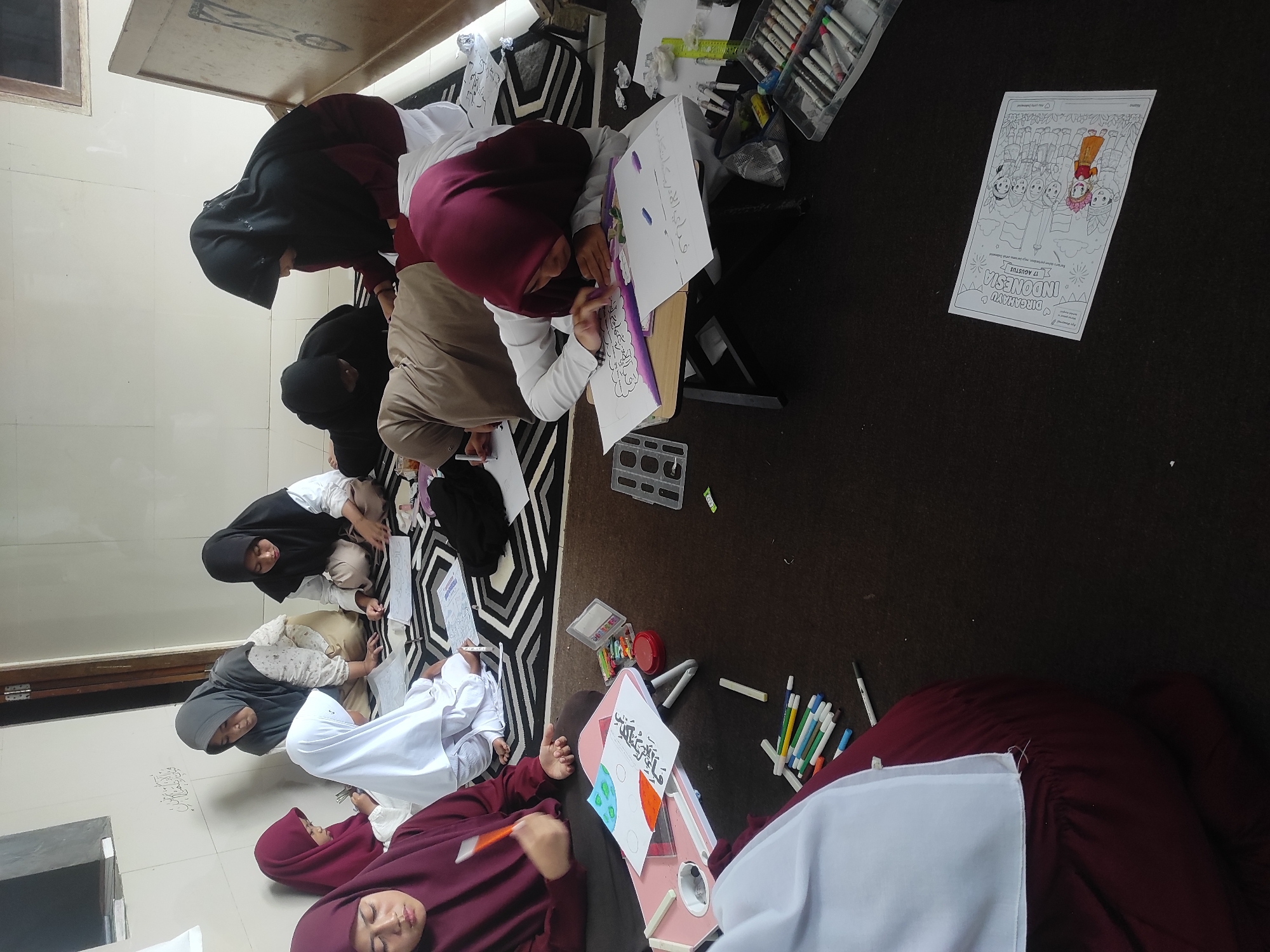 A group of at least seven young girls, dressed in hijabs and long-sleeved garments, are seated on the floor in a room, engaged in an art activity. The floor is covered by a dark brown carpet, with a black and white patterned rug underneath the seated children. The room appears to be indoors, with plain white walls. In the background, a wooden door frame is visible.
The girls are working on various drawing and coloring activities. Several of them are hunched over papers spread out on the floor or on small lap desks. Art supplies, including crayons, markers, pens, and rulers, are scattered around them. One girl in the foreground, wearing a maroon hijab and a matching maroon top, is holding an orange crayon and appears to be coloring. Another girl, in a white hijab, is also actively drawing. A girl in a black hijab is looking intently at her work, as are others.
The overall impression is one of focused creative activity in a communal setting. The presence of a coloring sheet with the text "DIRGAHAYU INDONESIA 17 AGUSTUS" suggests a patriotic theme to their art project, likely in commemoration of Indonesian Independence Day. The lighting in the room appears to be natural, coming from an unseen source, and casts soft shadows. The room is uncluttered, with the primary focus being on the girls and their art supplies. While the image does not explicitly state the location, the mention of "Indonesia" on the coloring sheet strongly suggests the activity is taking place within Indonesia. Given the mention of Sumedang, it is reasonable to infer that this scene is occurring in a school, community center, or home within that city.