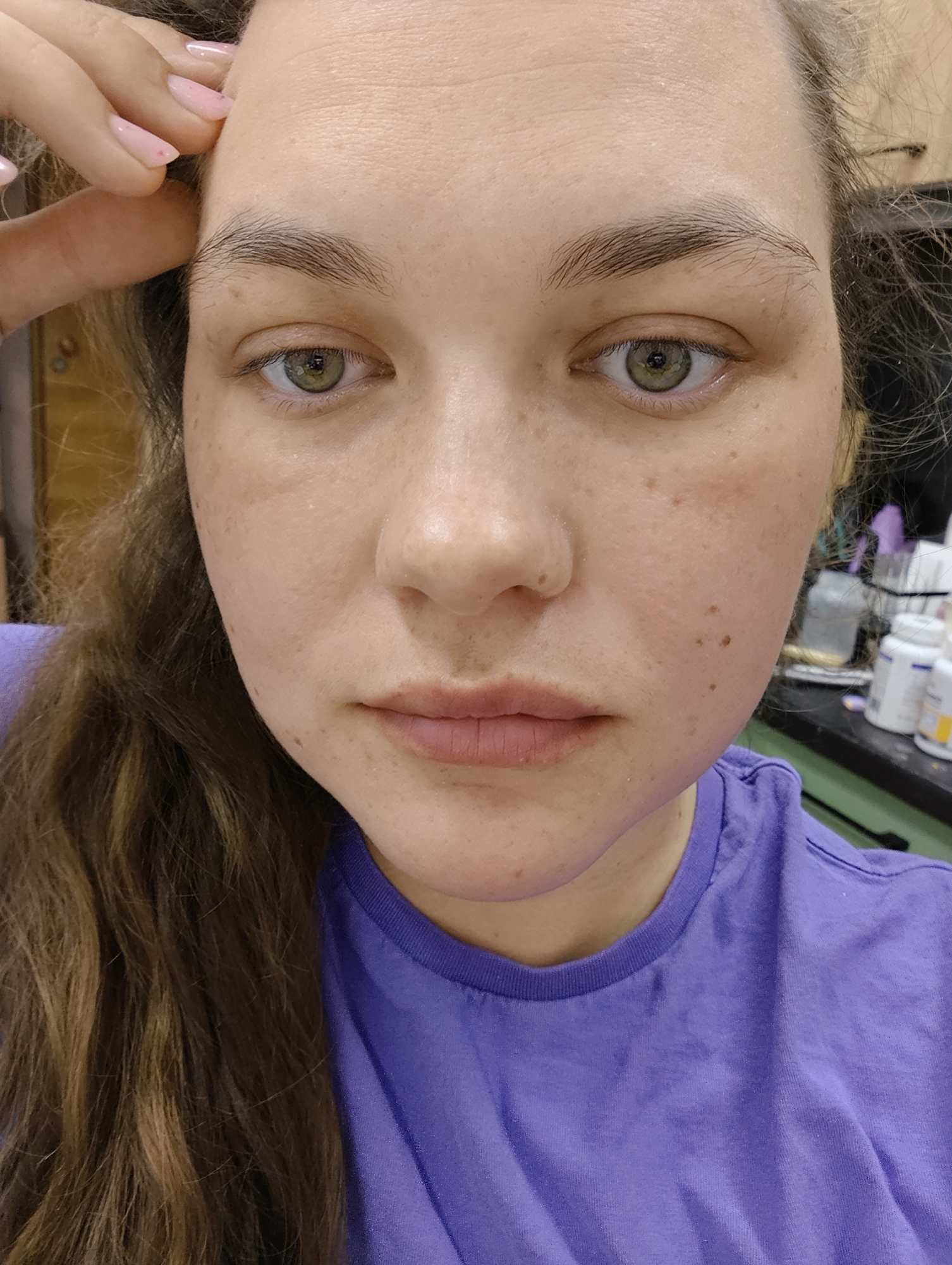 A close-up selfie of a Caucasian woman with fair skin, freckles, and light brown hair. She is wearing a purple t-shirt. Her eyes are green and her eyebrows are dark brown. Her right hand is raised to her forehead. The background is slightly blurred, showing a dark counter with various bottles and objects. The location is not discernible from the provided image. There is no evidence to indicate this photo was taken in Chaikovsky, Russia.