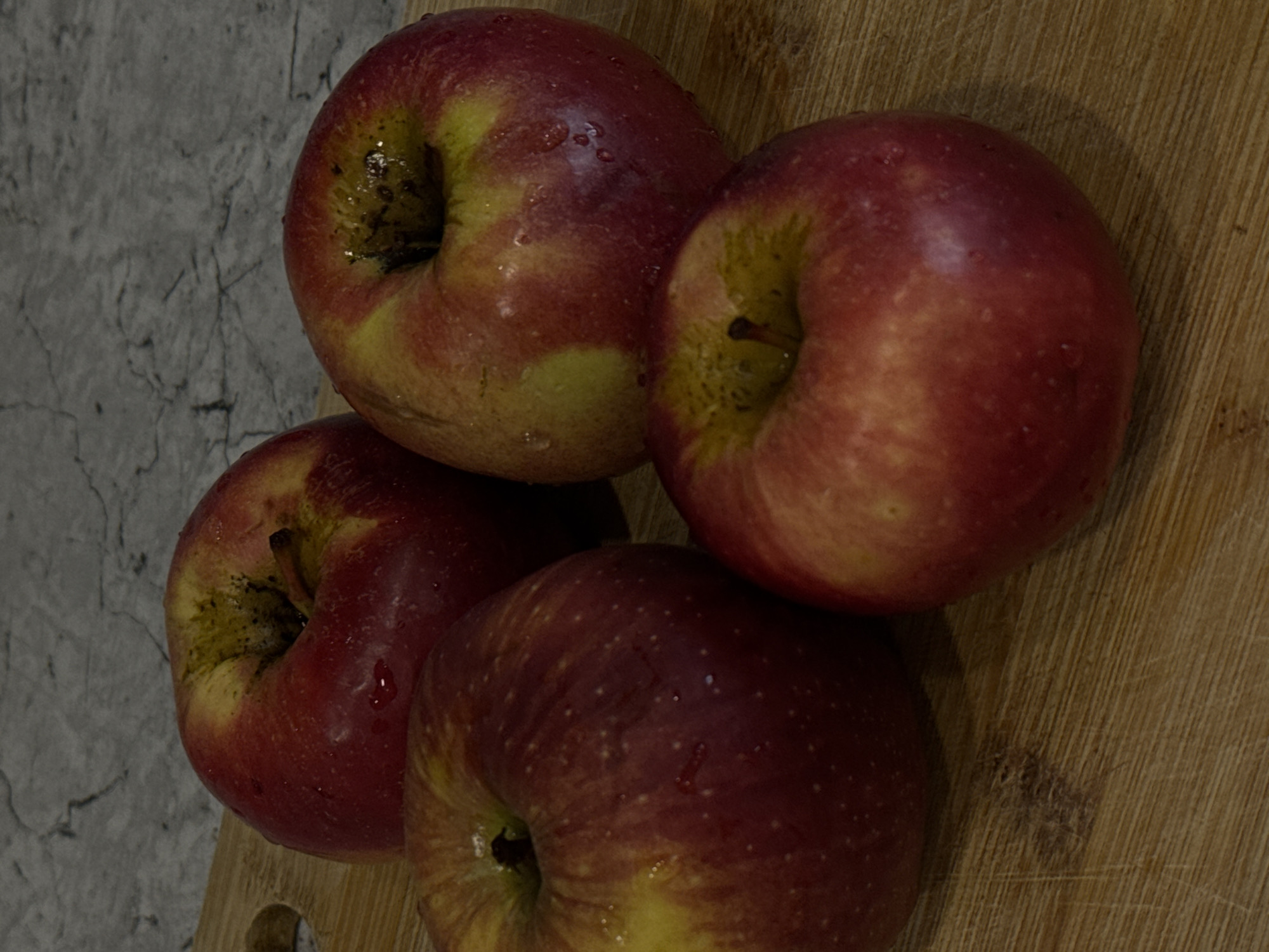 Four red and yellow apples are depicted in a still life, arranged on a wooden surface adjacent to a grey, patterned background. The primary focus is on the apples, which are clustered together. Each apple exhibits a mix of vibrant red and softer yellow hues, with some natural blemishes and variations in skin texture. Notably, there are visible water droplets on the surface of the apples, suggesting they may have been recently washed or are fresh.
The wooden surface, likely a cutting board, features a distinct grain pattern and appears to be lightly worn. In one of the cropped images, a circular cut-out, possibly part of a handle or hanging hole, is visible on this wooden surface. The grey background has a marble-like or concrete-like appearance, characterized by intricate dark veins and patterns.
The lighting in the image is somewhat dim, creating soft shadows that add depth to the composition, suggesting an indoor setting under artificial light. No specific activity or event is taking place, and there is no visible text. The image captures the texture and color of the fruit and the contrasting surfaces it rests upon.