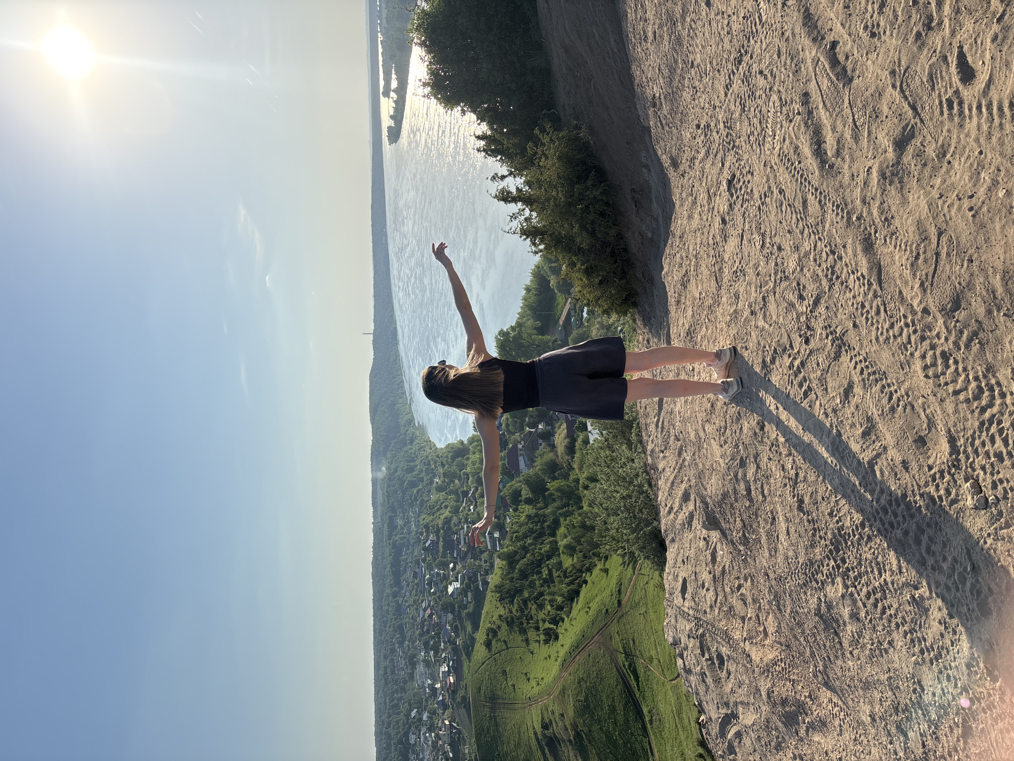A woman stands on the edge of a cliff overlooking the river in Ольховка, Russia. The cliff is sandy, and there are numerous footprints in the sand. The woman is wearing a dark top and skirt. She is standing with her arms outstretched, with one arm fully extended upwards. In the background, there is a river with greenery and buildings, trees visible on the right side of the river. In the foreground, on the left side, a village/small town is visible on a slope that leads up to the cliff edge. The sun is visible in the sky, producing a lens flare.