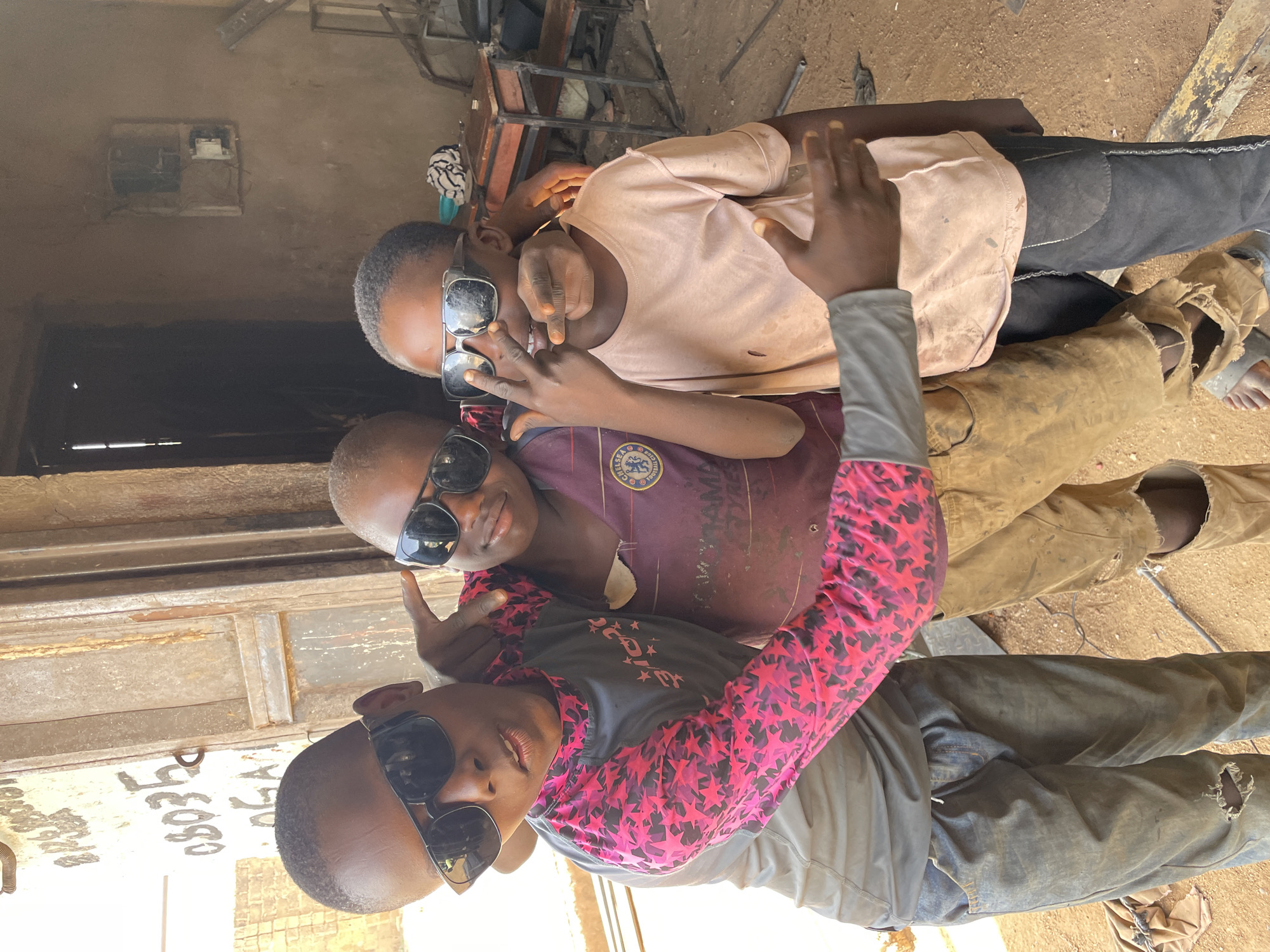 Three young boys are posed in front of what appears to be a storefront or shop in Barakin Akawo, Nigeria. All three are wearing sunglasses. The boy at the bottom of the frame wears a black and pink star-patterned long-sleeved shirt with a gray jacket. The boy in the middle is wearing a dark-colored soccer jersey with the Chelsea FC logo visible. The boy at the top is wearing a light-colored t-shirt. They appear to be posing for a photo together. The environment is somewhat cluttered, possibly an industrial or commercial area, with building materials and metal structures visible. The storefront behind them has peeling paint and illegible text. The ground appears to be a mix of dirt and concrete.