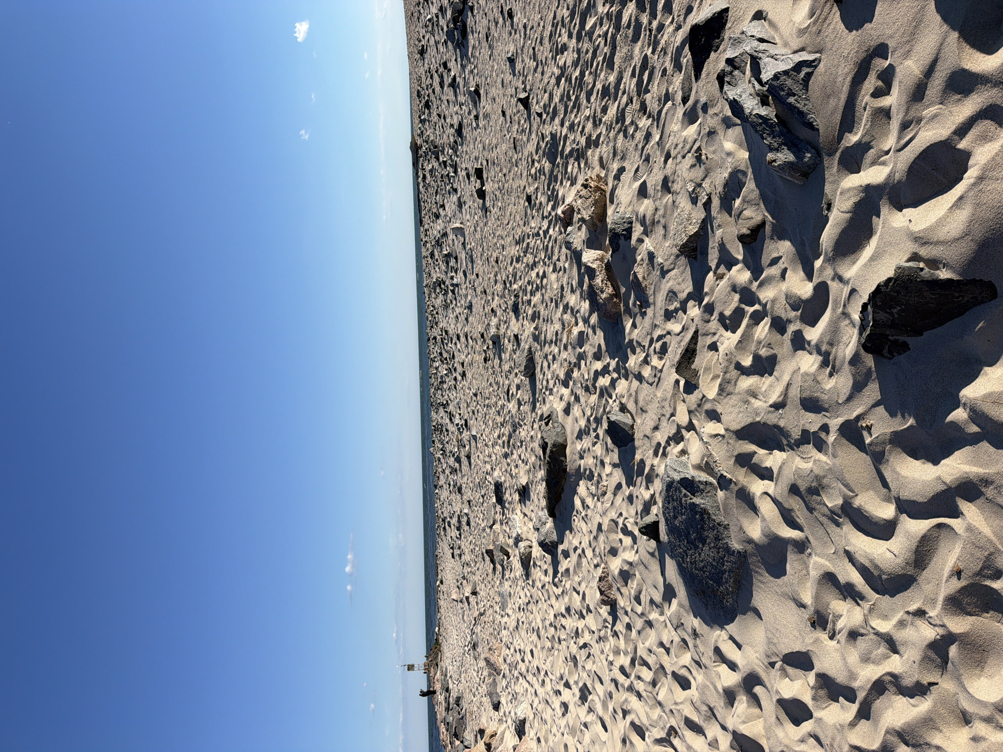 The image depicts a wide, sandy beach with numerous rocks scattered across the surface. The sand is textured with footprints and wave-like patterns. In the background, the ocean stretches to the horizon under a clear blue sky with a few wispy clouds. On the left side of the image, a rocky pier extends into the water, with two individuals standing near a navigational marker. The scene is bathed in sunlight, casting long shadows across the sand. The location is identified as a beach in Hampton Bays, United States of America.