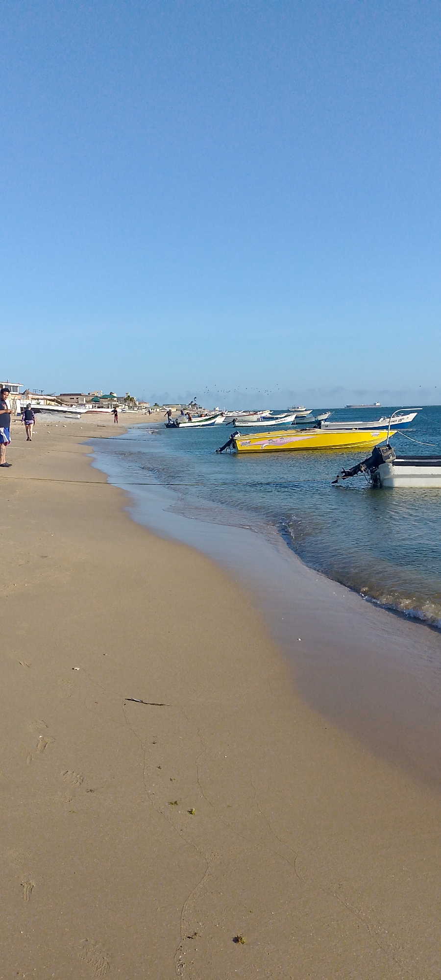 The image captures a beach scene in Villa Marina, Venezuela. The foreground is dominated by a sandy beach with a visible waterline. Several boats are moored in the shallow water, including a distinct yellow boat. People are walking along the shoreline in the background. The sky is clear and blue. A few buildings are visible in the far background, near the horizon.