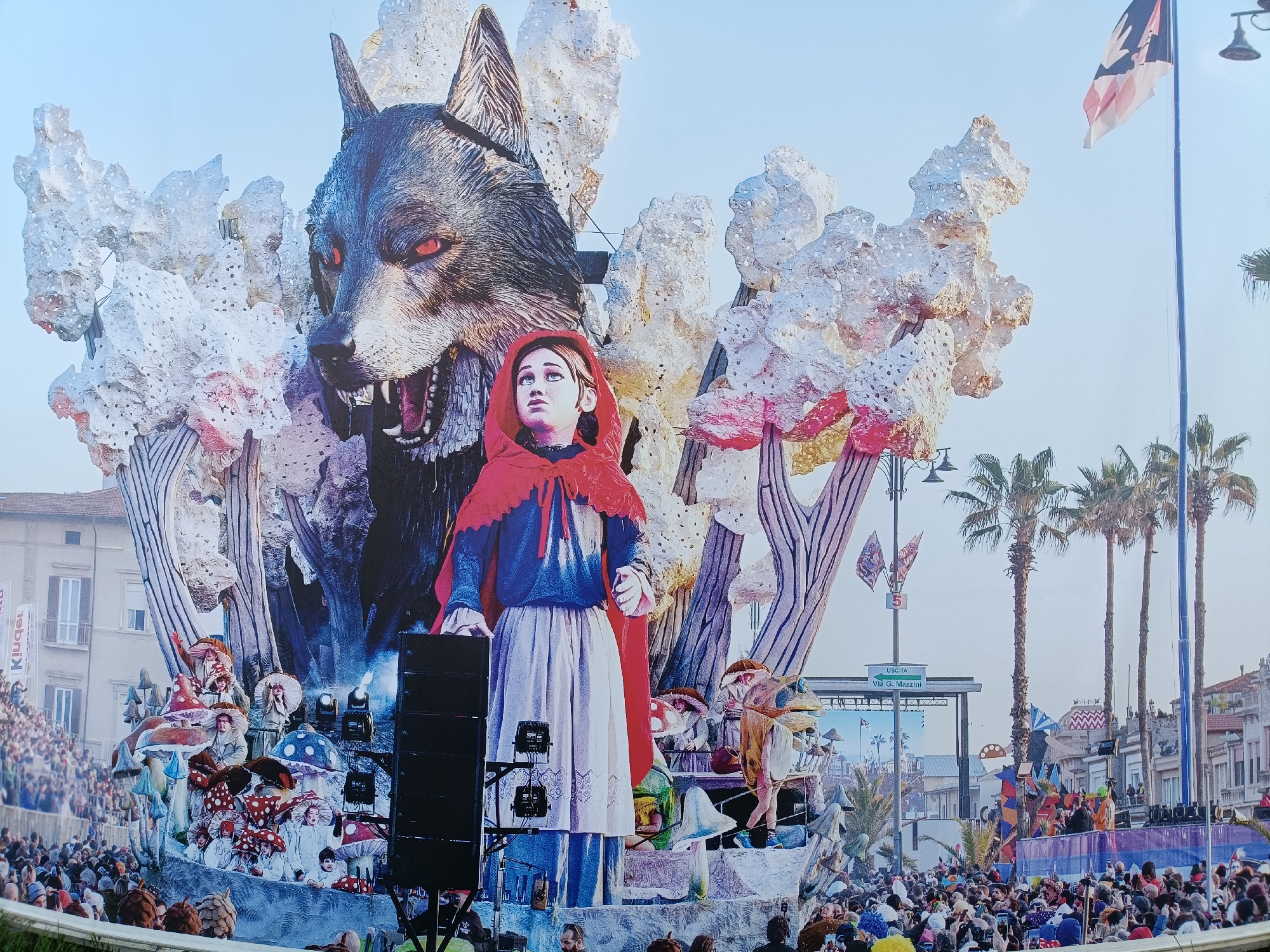 The image captures a vibrant carnival float parade in Viareggio, Italy, likely during the annual Viareggio Carnival. The focal point is a colossal float depicting a scene from a fairy tale. Dominating the upper portion is a massive, menacing wolf's head with glowing red eyes and bared teeth, appearing to loom over a young girl dressed as Little Red Riding Hood, complete with a red cape. The float is adorned with large, abstract, snow-like or cloud-like structures, some tinged with pink.
Below the main characters, the float is populated by performers dressed as fantastical mushroom-like figures, some with large hats, and other whimsical characters in costume. The base of the float is decorated with colorful mushrooms of various sizes. In front of the float, a large black speaker system is positioned.
The parade is taking place on a street lined with buildings, palm trees, and streetlights. A crowd of spectators is visible on the sides, some standing behind a barrier in the foreground, indicating a public event. Flags, including one with a black and white emblem, are visible, and a street sign indicating "USCITA Via G. Mazzini" (Exit Via G. Mazzini) suggests the parade route or location within the city. The sky is a clear blue. The overall impression is one of a lively and elaborate festive occasion.