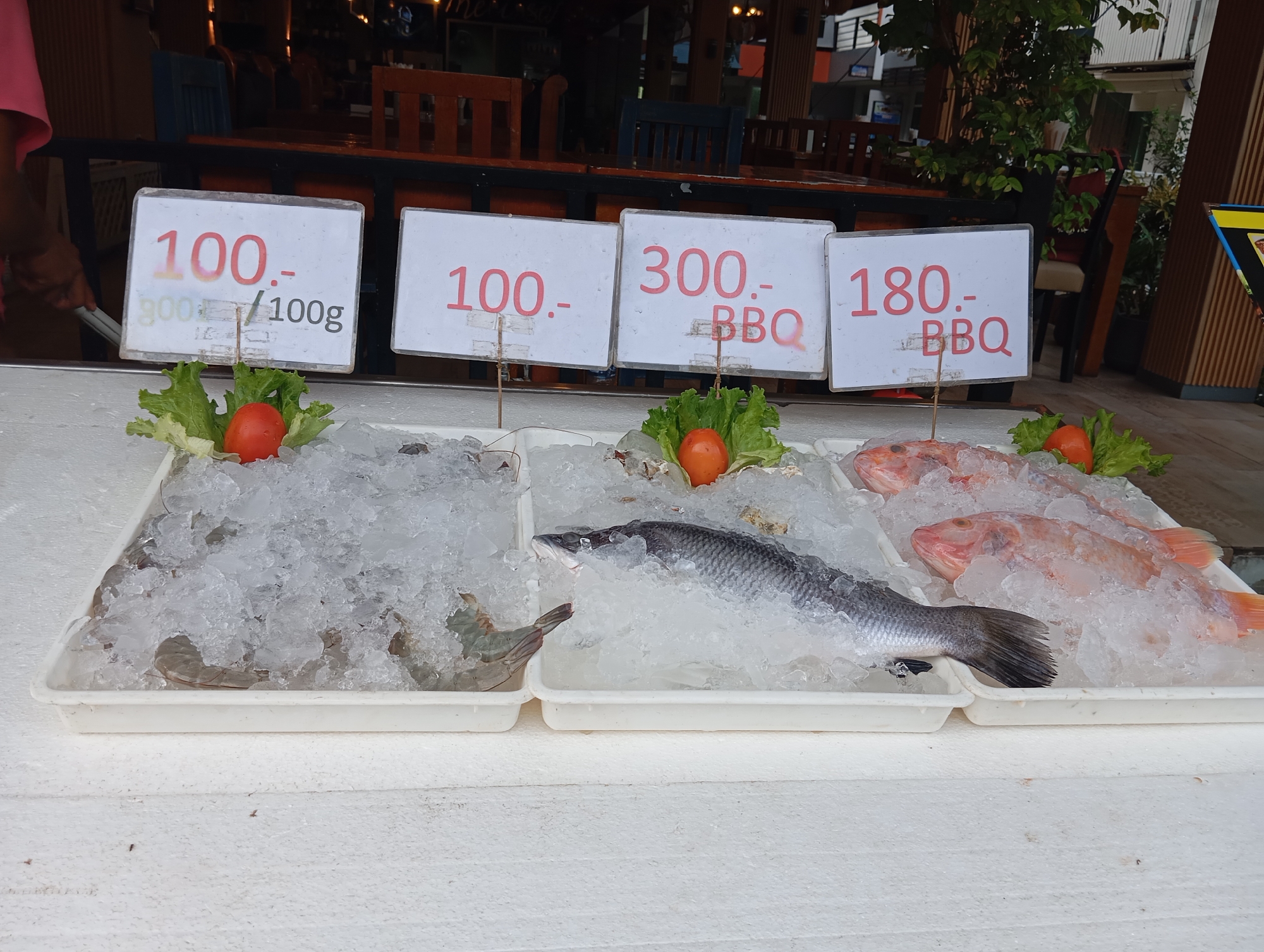 The image shows a seafood display on a table outdoors, likely in Karon, Thailand, given the context. In the foreground, three white plastic trays are filled with ice and seafood. The leftmost tray contains shrimp, partially obscured by ice. The middle tray holds a whole fish, likely a barramundi, and some shrimp. The rightmost tray contains several red-skinned fish, possibly snapper. Each tray has a price sign in front of it. The signs display prices in Thai Baht, with the leftmost sign showing "100.- / 100g", the middle sign "300.- BBQ", and the rightmost sign "180.- BBQ". Fresh garnishes of lettuce and tomatoes are placed on top of the ice in front of the signs. In the background, there are wooden tables and chairs, suggesting a restaurant or food stall setting. A person's arm, wearing a pink shirt, is visible on the far left, and a hint of outdoor foliage is present on the right. The overall scene depicts a fresh seafood market display.