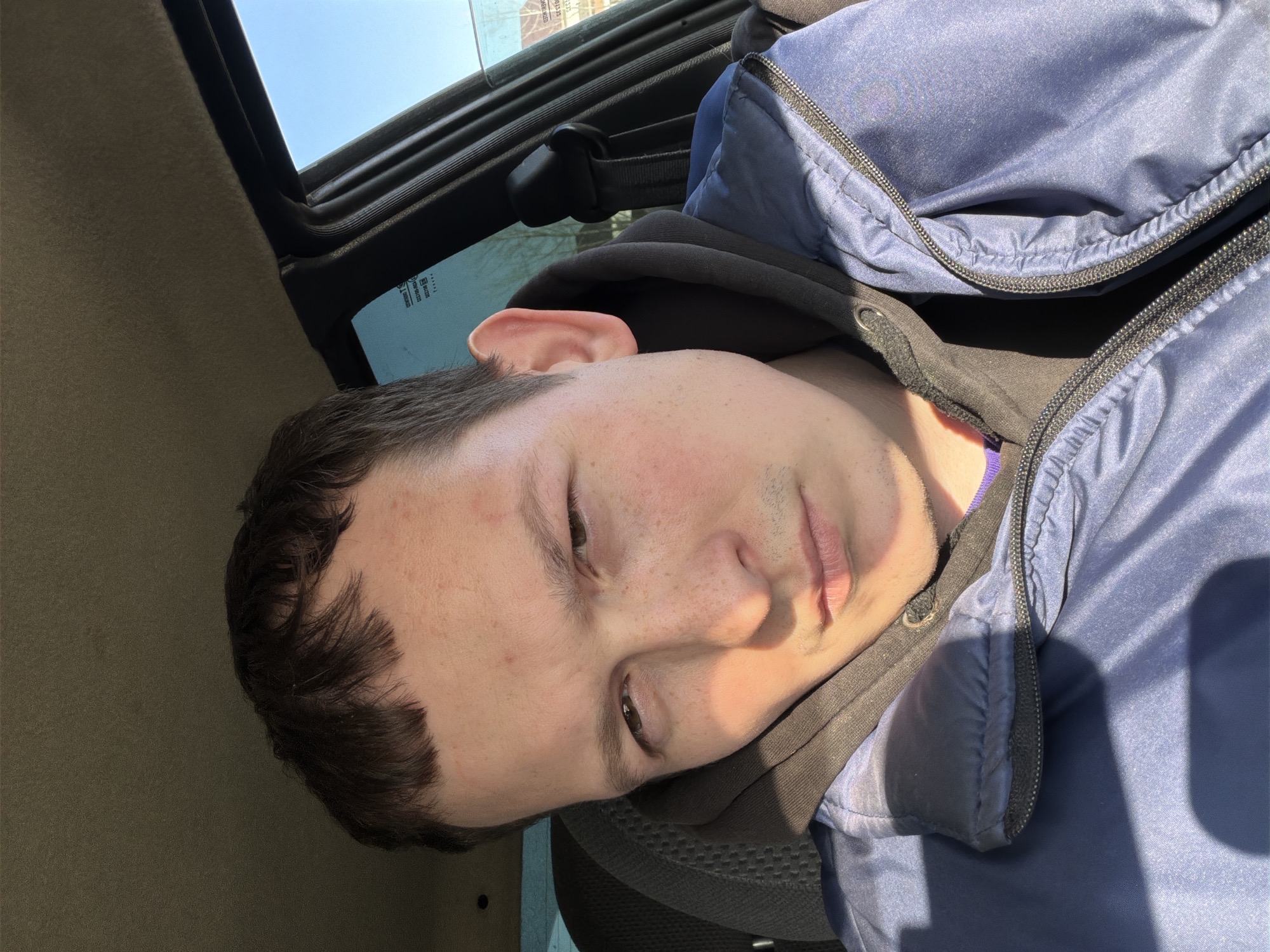 A man is photographed in the front seat of a vehicle. He is wearing a blue jacket over a black hooded sweatshirt. The vehicle's sunroof is open. The location cannot be confirmed as Прикамский, Russia without additional information or context within the image.