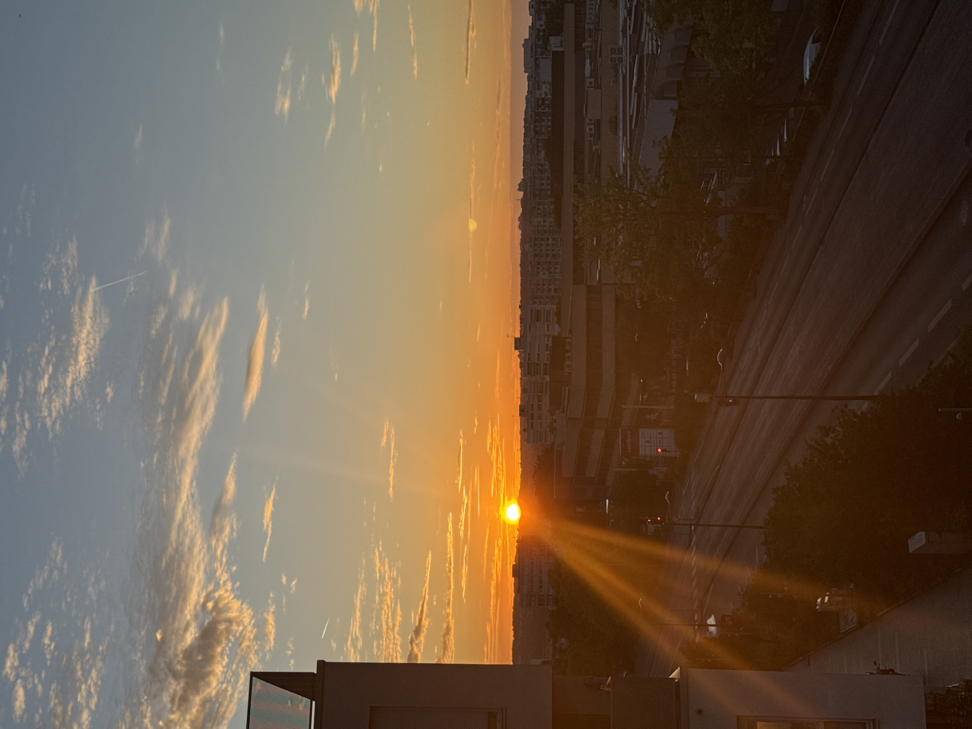 An aerial view captures a city skyline at sunset. The sun, a bright orb, is positioned just above the horizon, casting a warm orange glow across the sky. Sunbeams emanate from the sun, stretching across the scene. Wispy clouds are scattered in the sky, tinted with orange and pink hues.
In the foreground, a modern building with a glass facade is visible on the left. Below, a multi-lane road runs horizontally through the frame, bordered by trees and other urban structures. Traffic lights are visible along the road, some showing red. In the distance, a dense cluster of buildings forms the city's skyline, stretching across the horizon. The overall scene is bathed in the golden light of the setting sun, creating a serene and picturesque urban landscape.